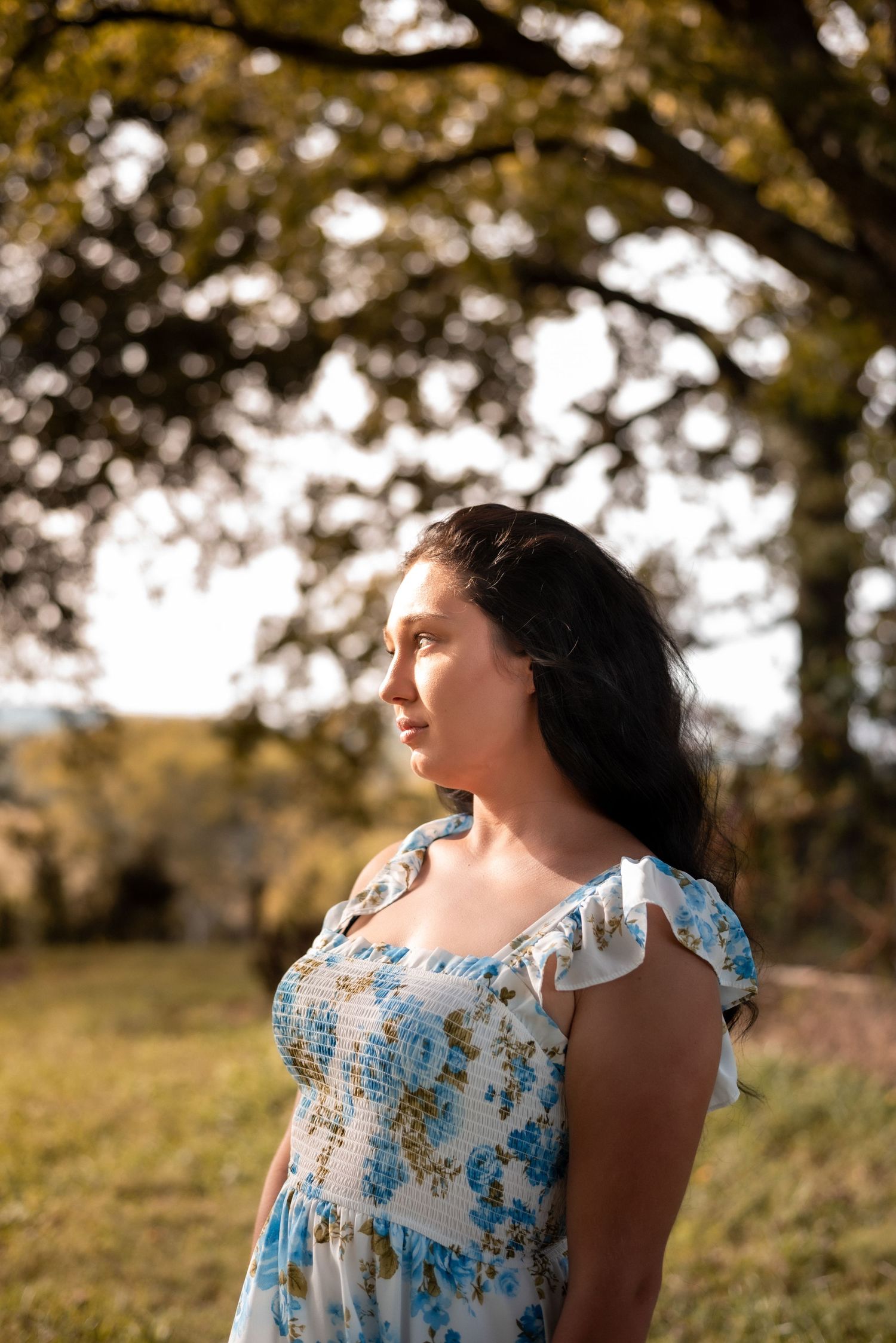 A person in a blue floral dress poses in front of sunlit trees during golden hour.