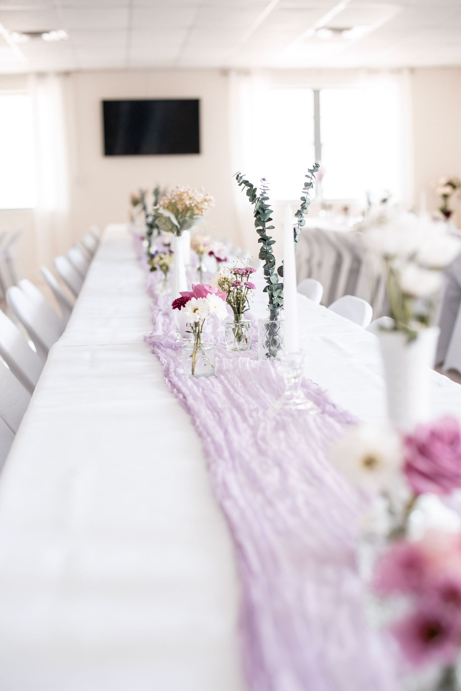 Elegant wedding reception table with lavender cheesecloth runner, small floral arrangements, and white chairs in a bright venue.