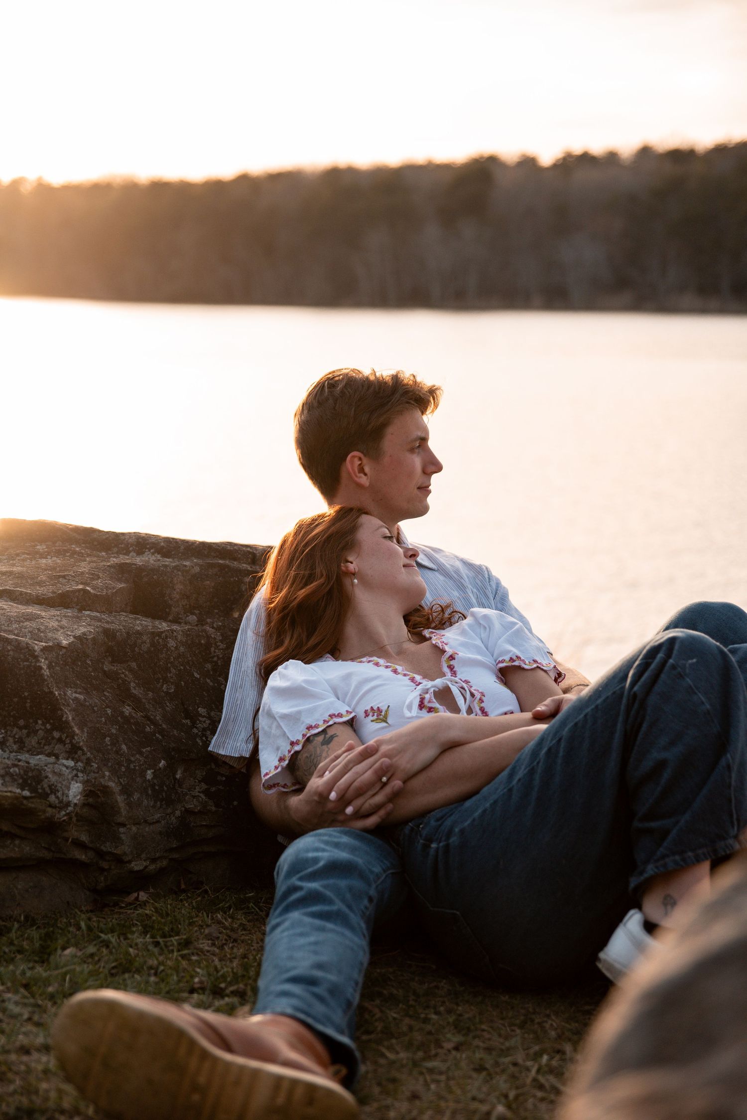Couple relaxing by a lake at sunset, woman leaning on man while he holds her gently on a grassy shore.