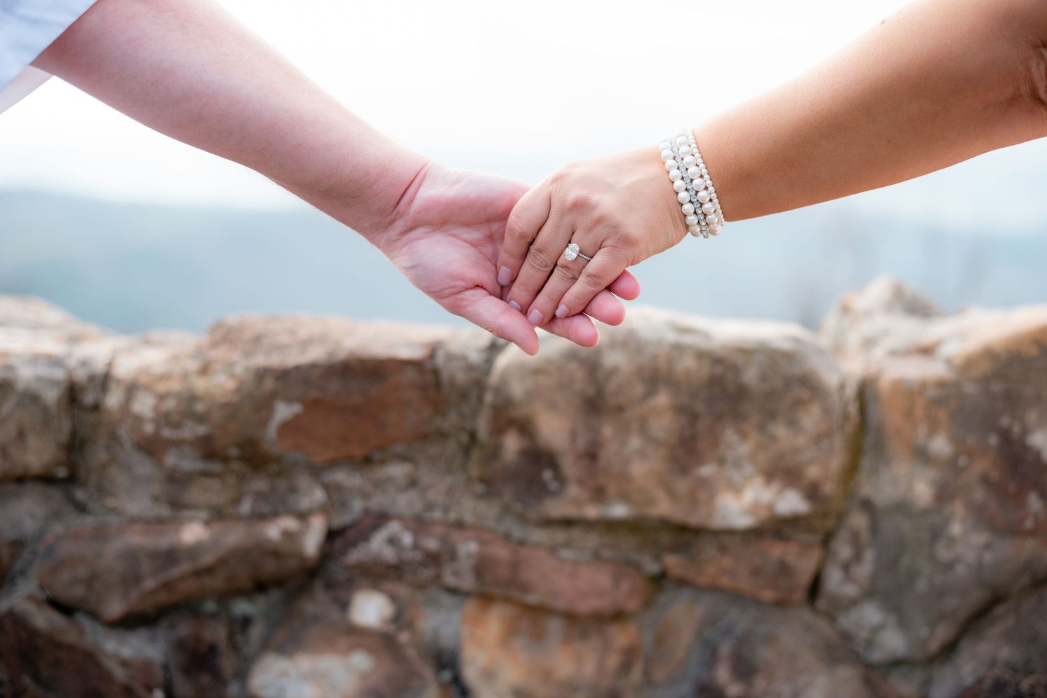 Two people holding hands outdoors near a stone wall, one wearing a diamond ring and pearl bracelet, symbolizing love and commitment.