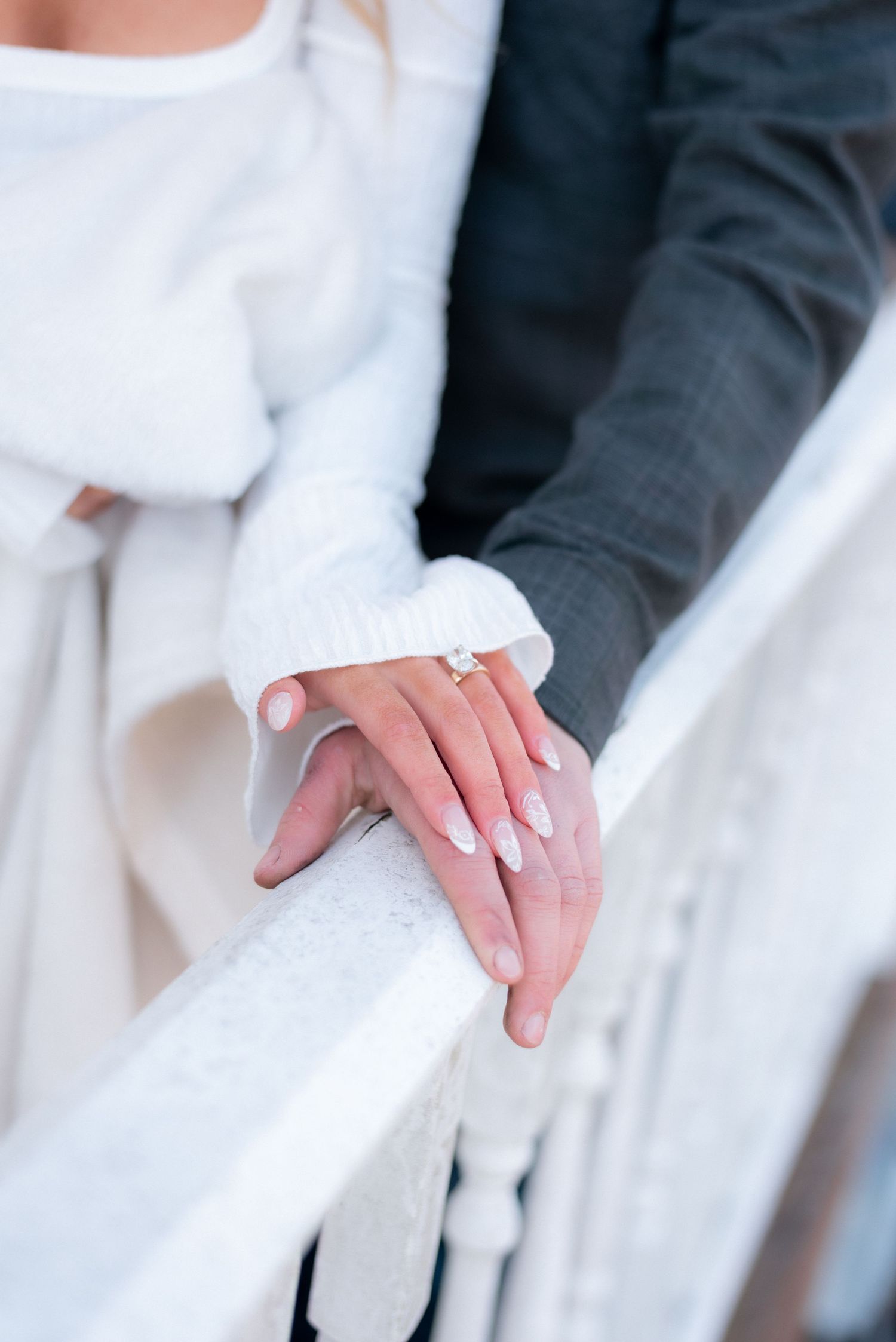 Close-up of hands with white manicured nails and engagement ring against formal attire.