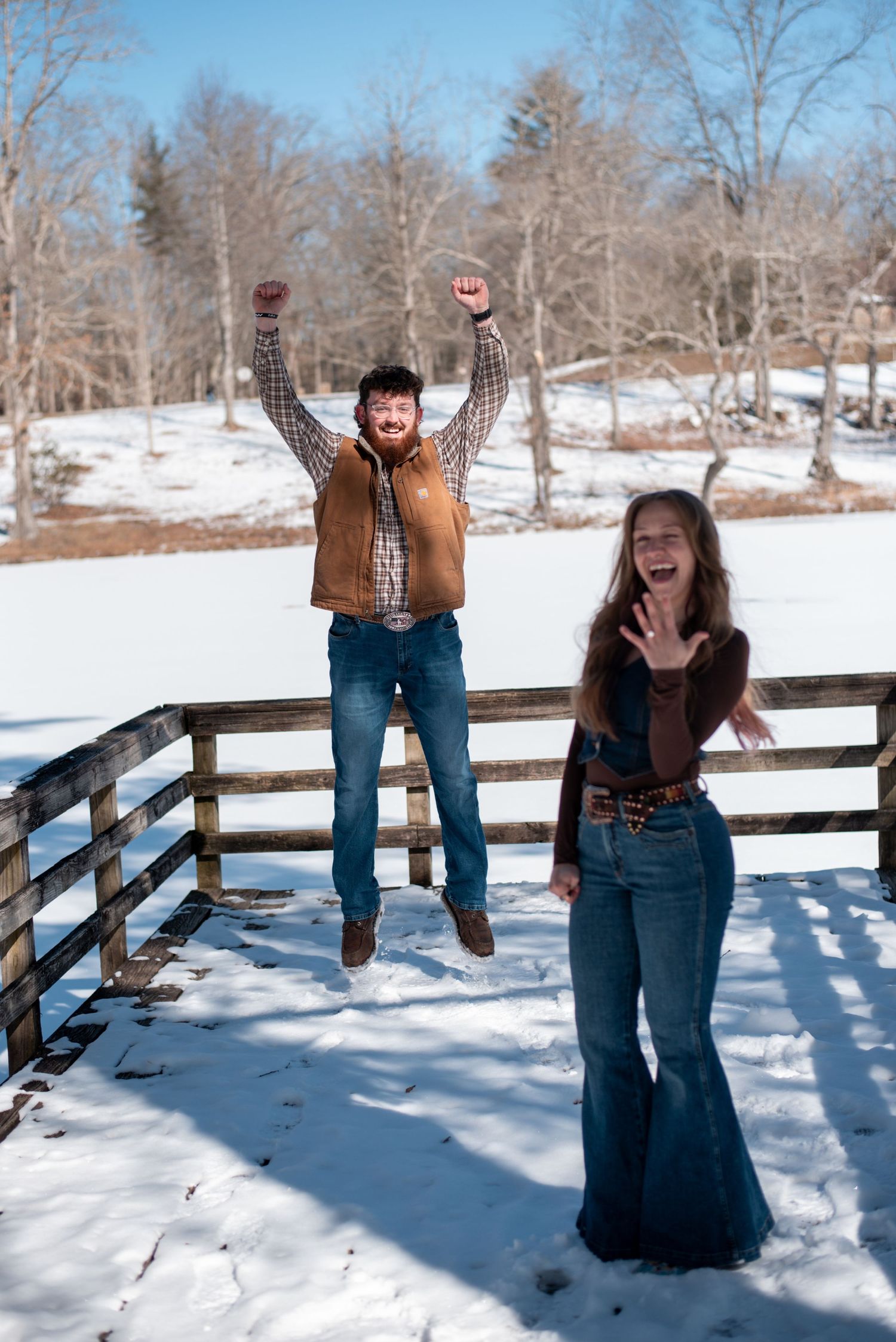 Two people celebrating joyfully on a snowy wooden deck with winter trees in the background.