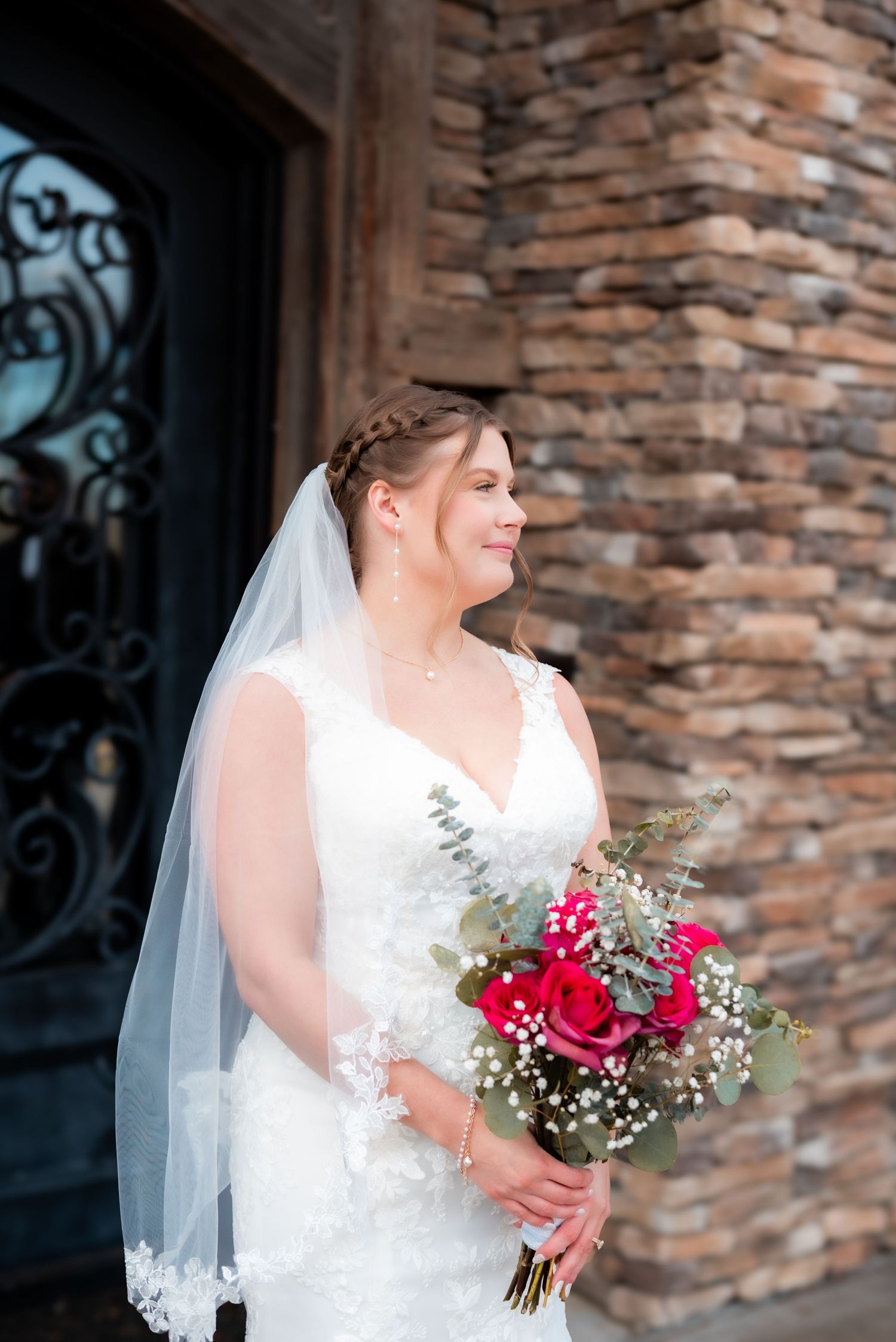 Bride in lace dress holds vibrant red and white bouquet against stone wall backdrop.