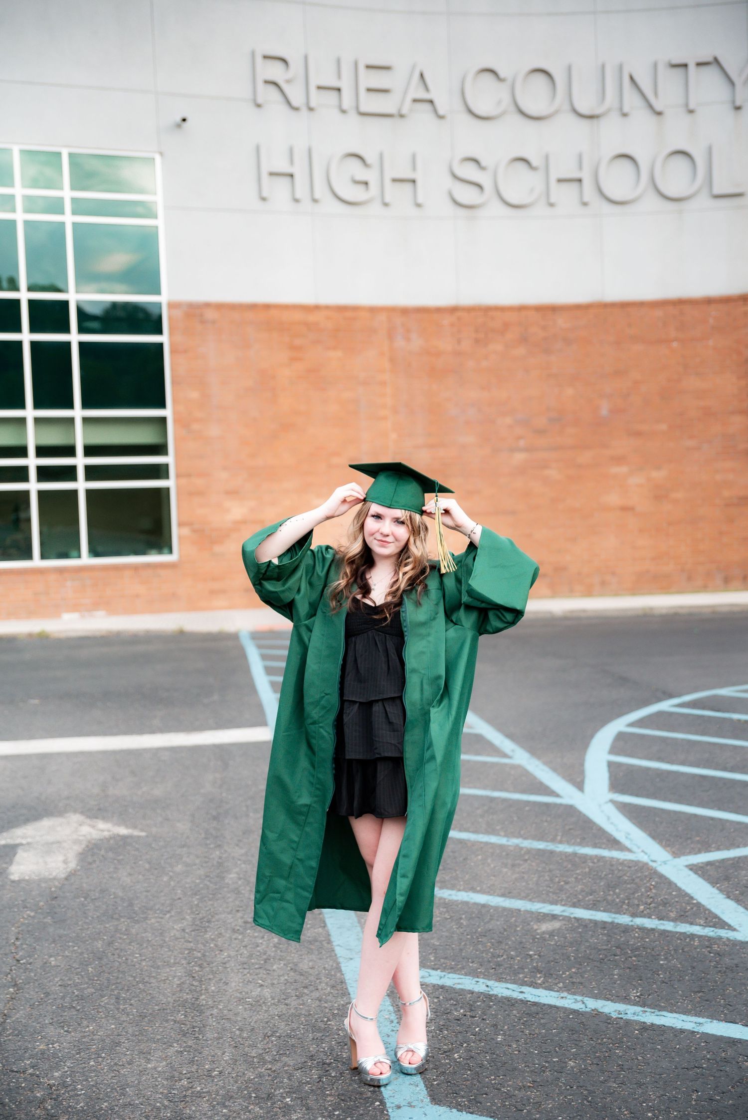 A smiling graduate in green cap and gown poses proudly in front of Rhea County High School.