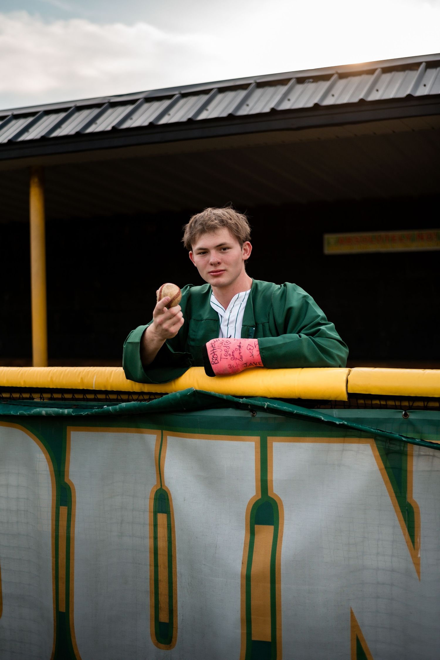 Young man in green jacket holds baseball at stadium dugout railing, wearing pink wristband on cloudy day.