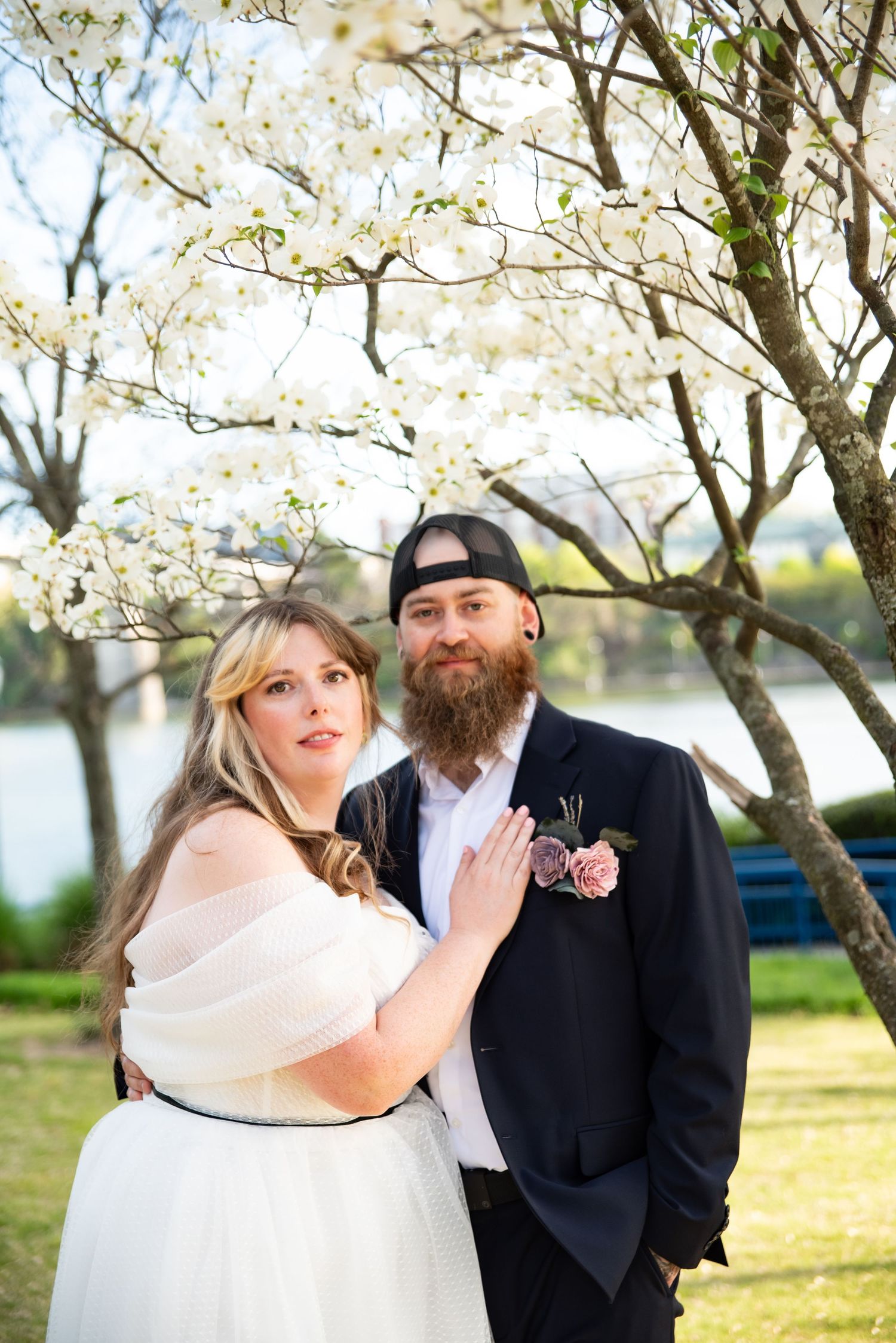 Bride and bearded groom in black suit pose under blooming white dogwood tree near a lake on their wedding day.