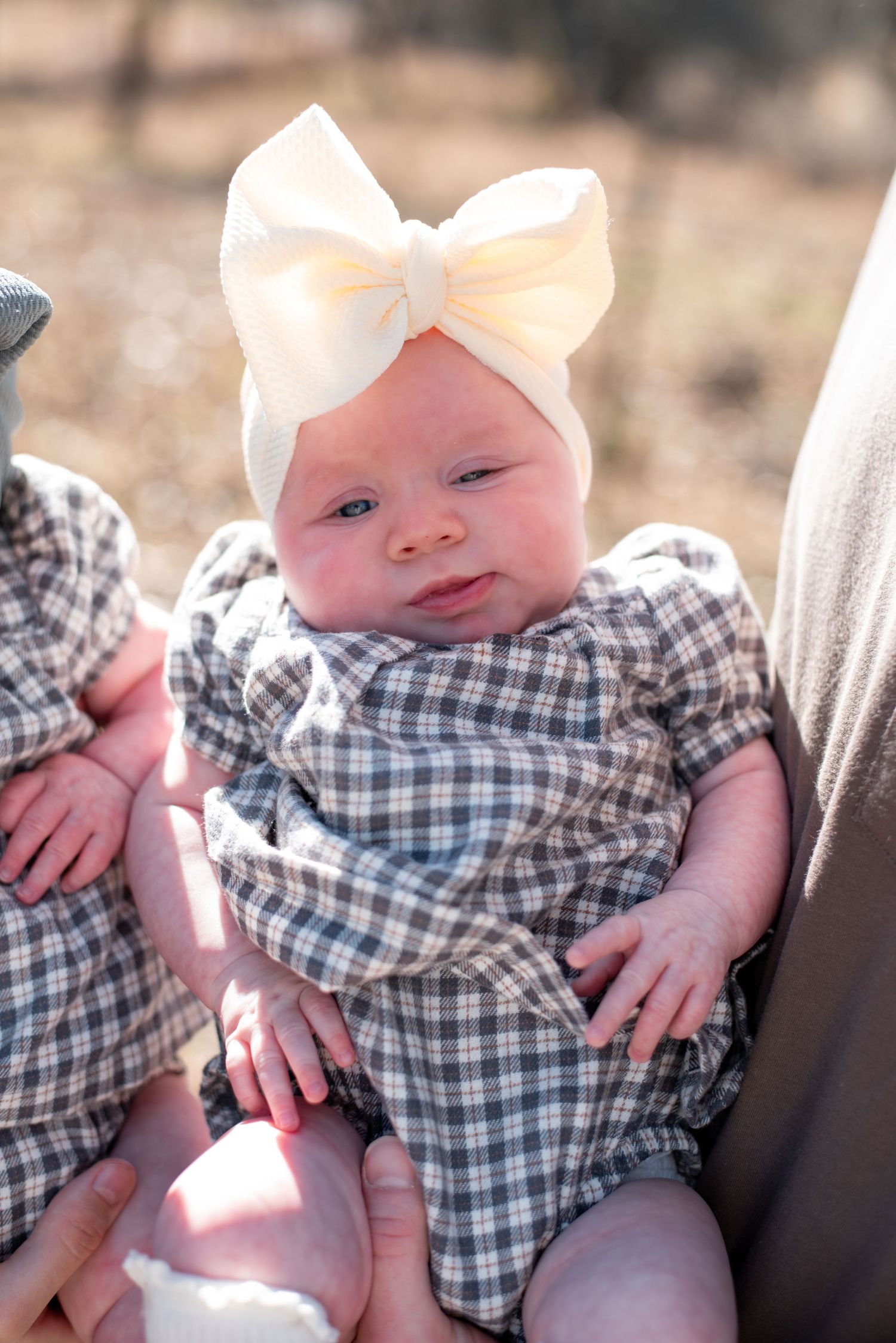 Baby in checkered dress with white bow smiling in outdoor portrait series.