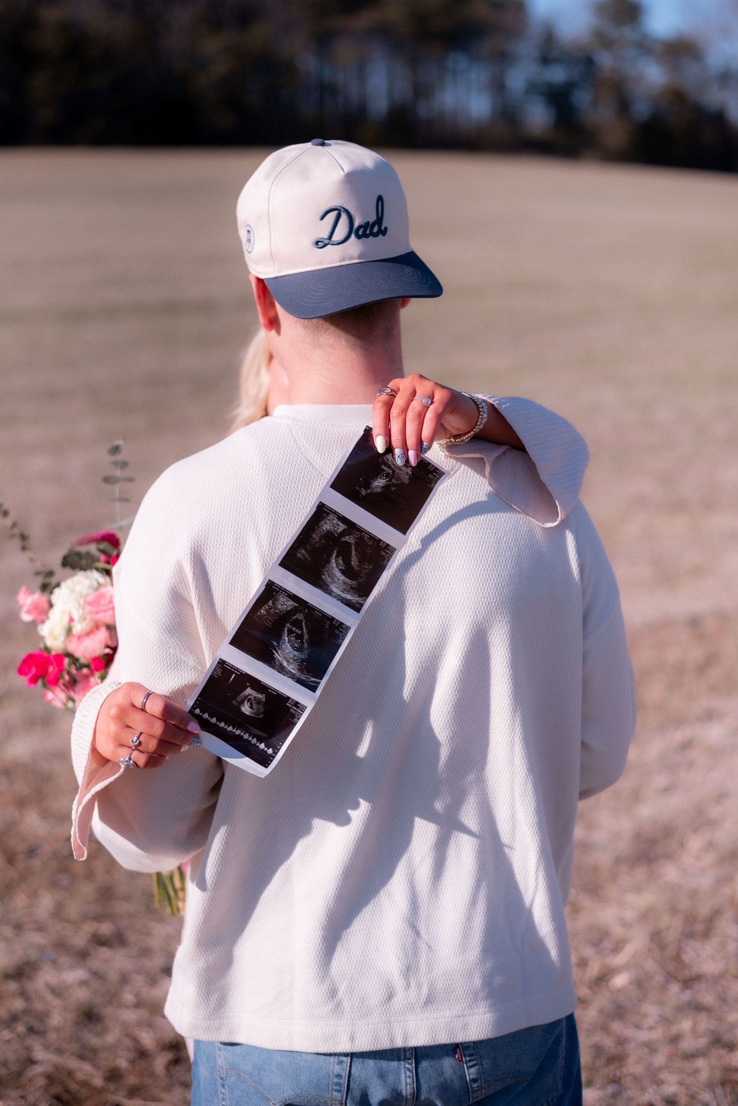 Someone wearing a Dad hat holds ultrasound photos and a bouquet against a natural backdrop.