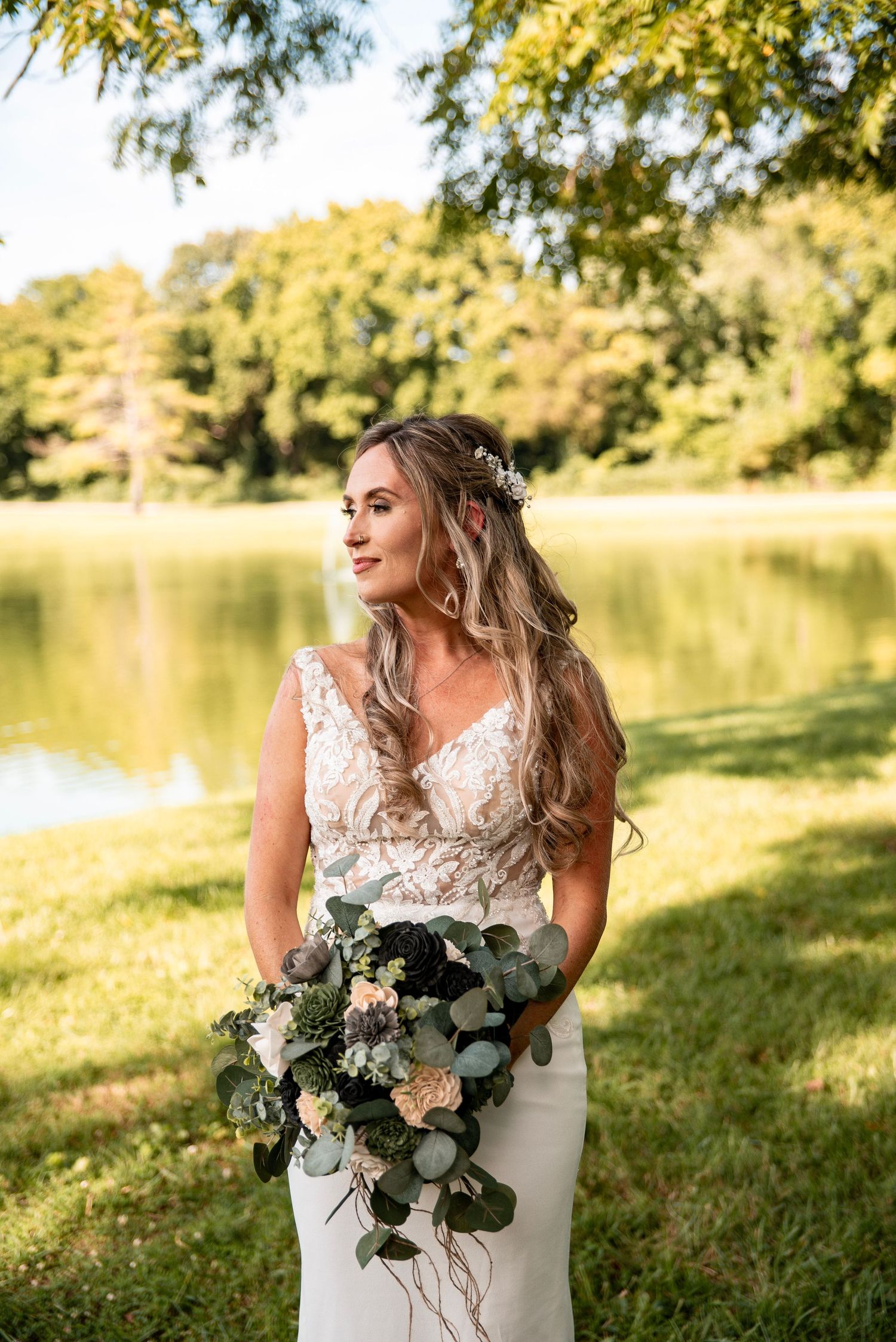 Bride in lace gown holding greenery bouquet stands by a sunlit pond surrounded by lush trees on her wedding day.
