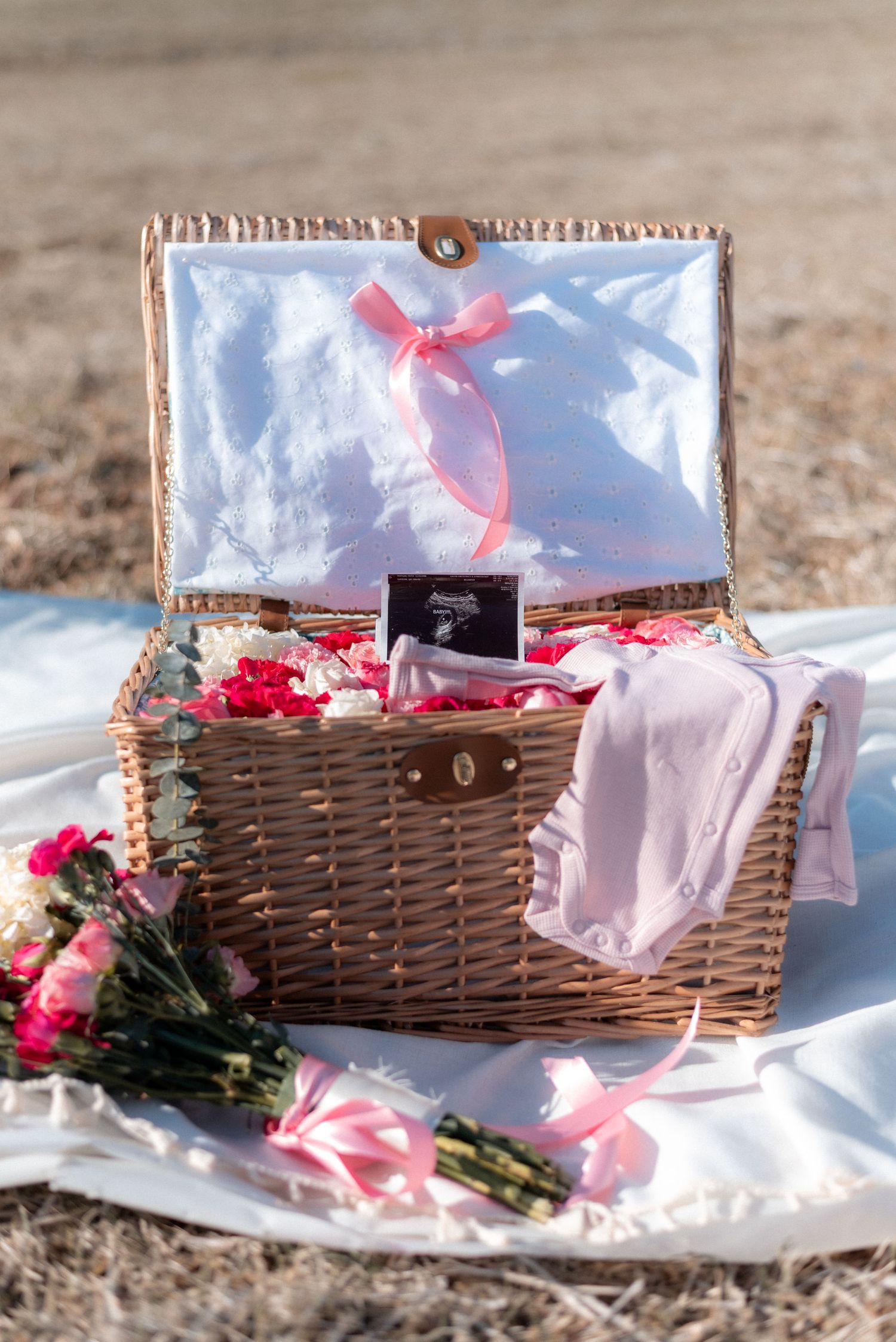 Romantic picnic setup with wicker basket, flowers and pink accents on white blanket.
