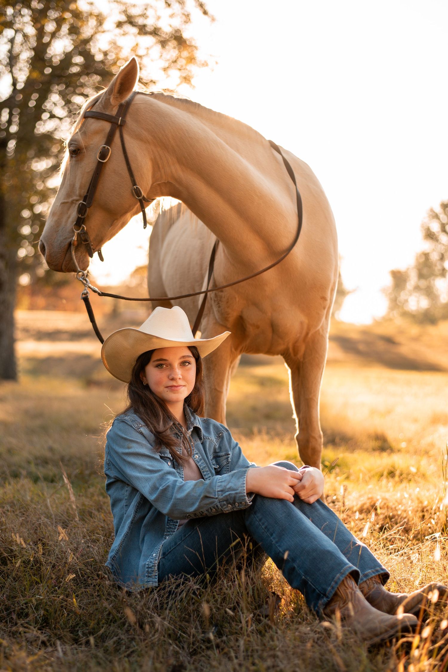 A person in western attire sits beside a palomino horse during golden hour.