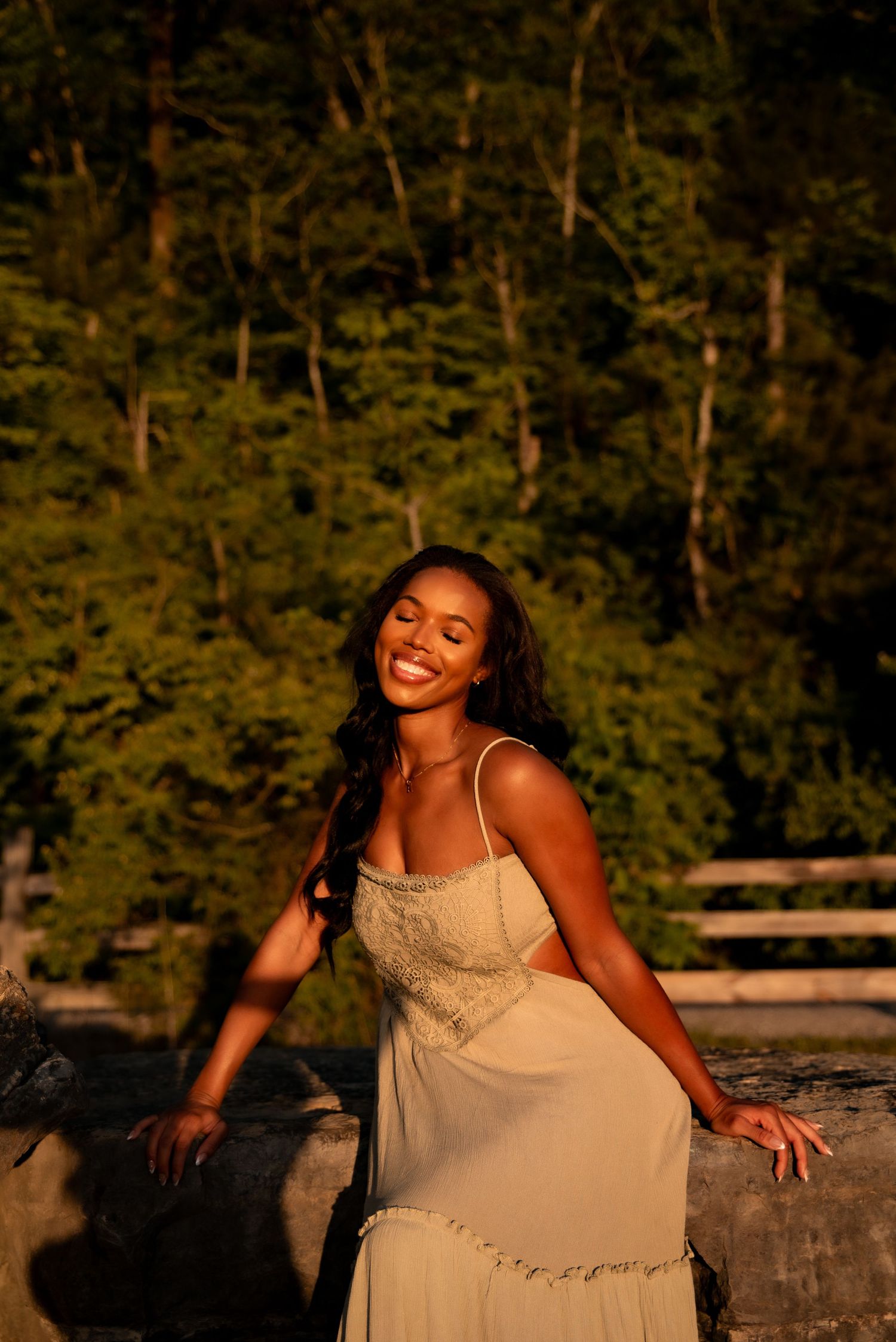 Joyful woman in cream lace dress smiling at golden sunset near stone wall outdoors.