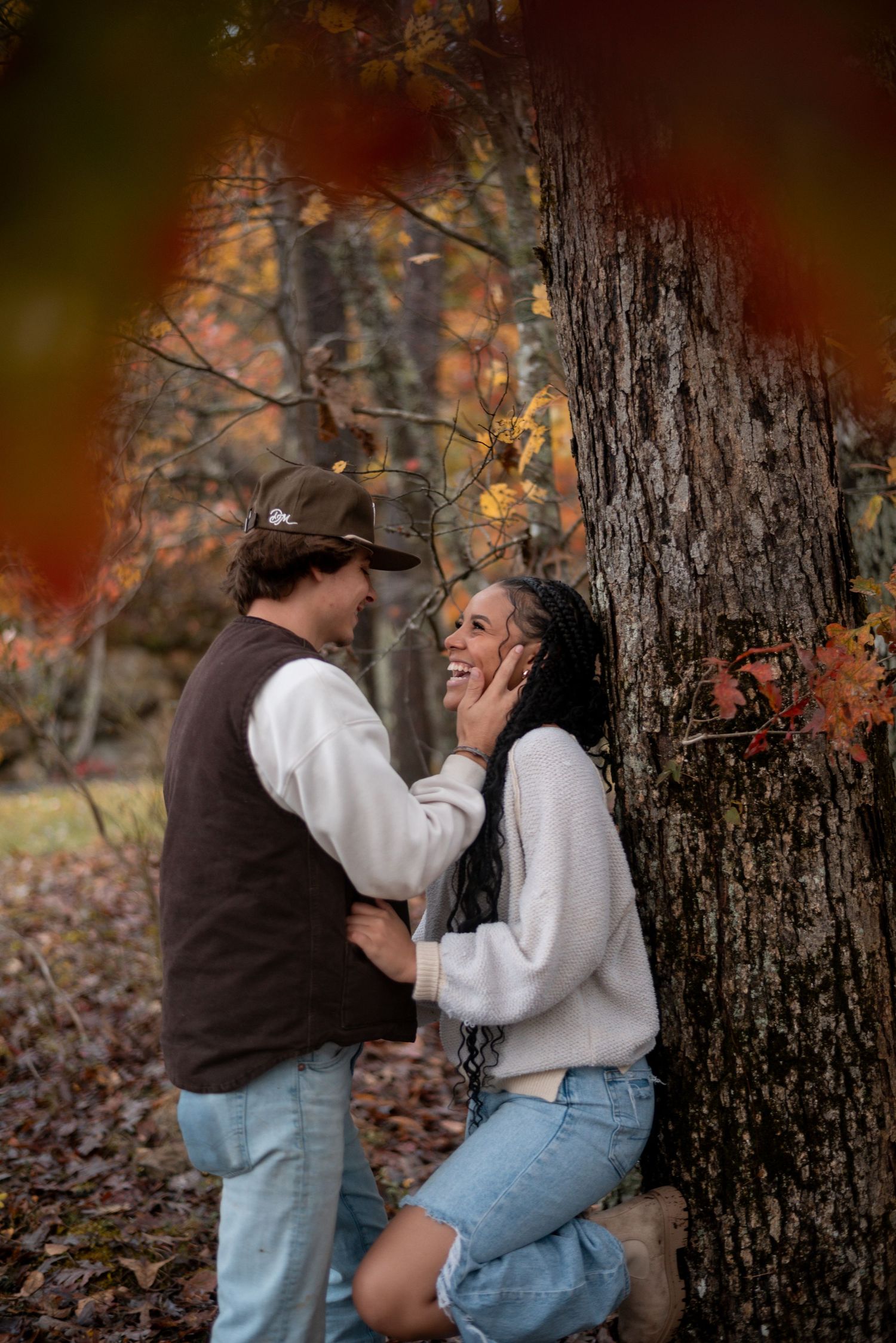 A romantic moment unfolds between two people by a large tree trunk in a fall forest setting.