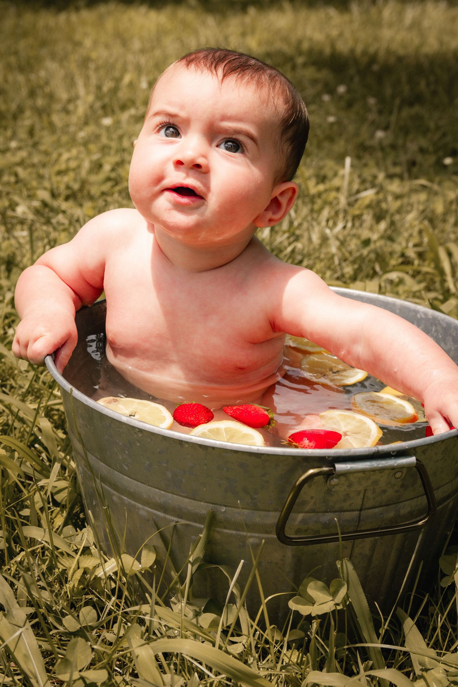 Baby taking bath in vintage metal tub with citrus slices and flowers.