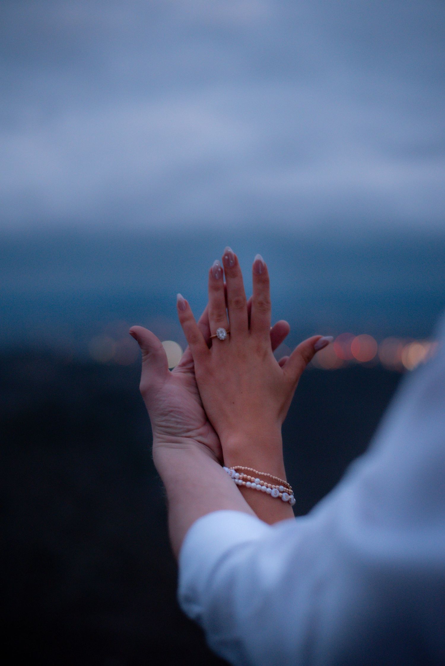 Two hands touching at dusk, one wearing a diamond engagement ring and pearl bracelet, with city lights bokeh background.