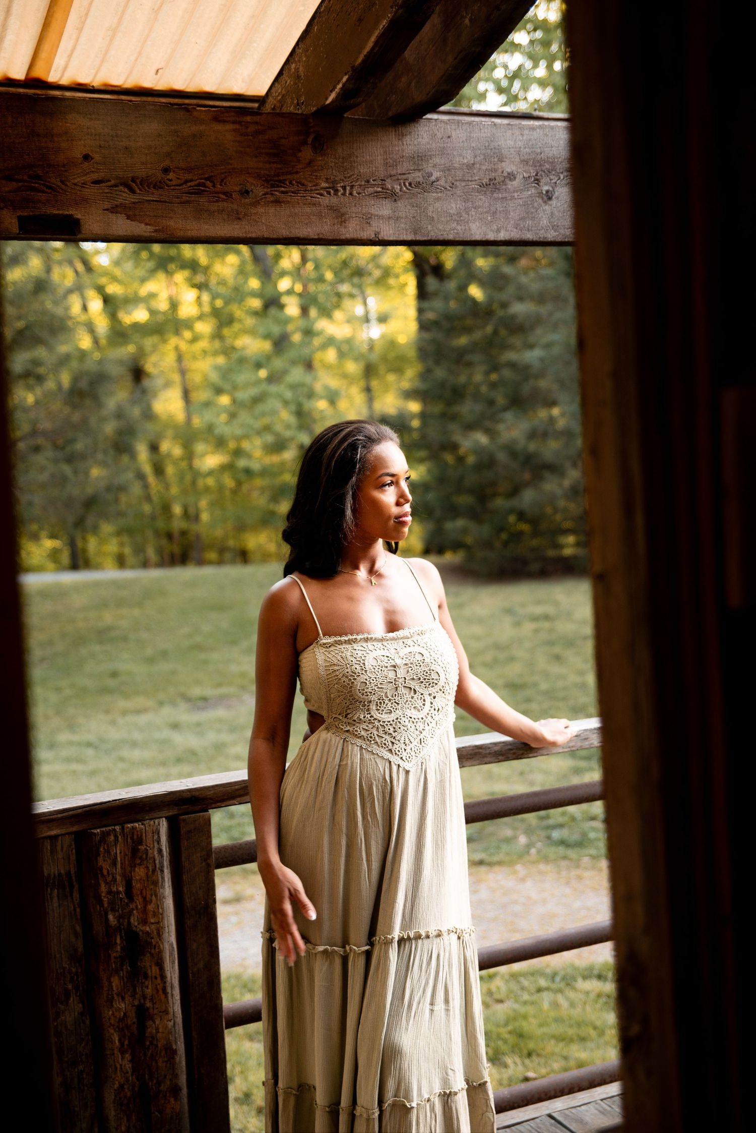 Young woman in lace cream sundress gazing outdoors from rustic wooden barn porch.