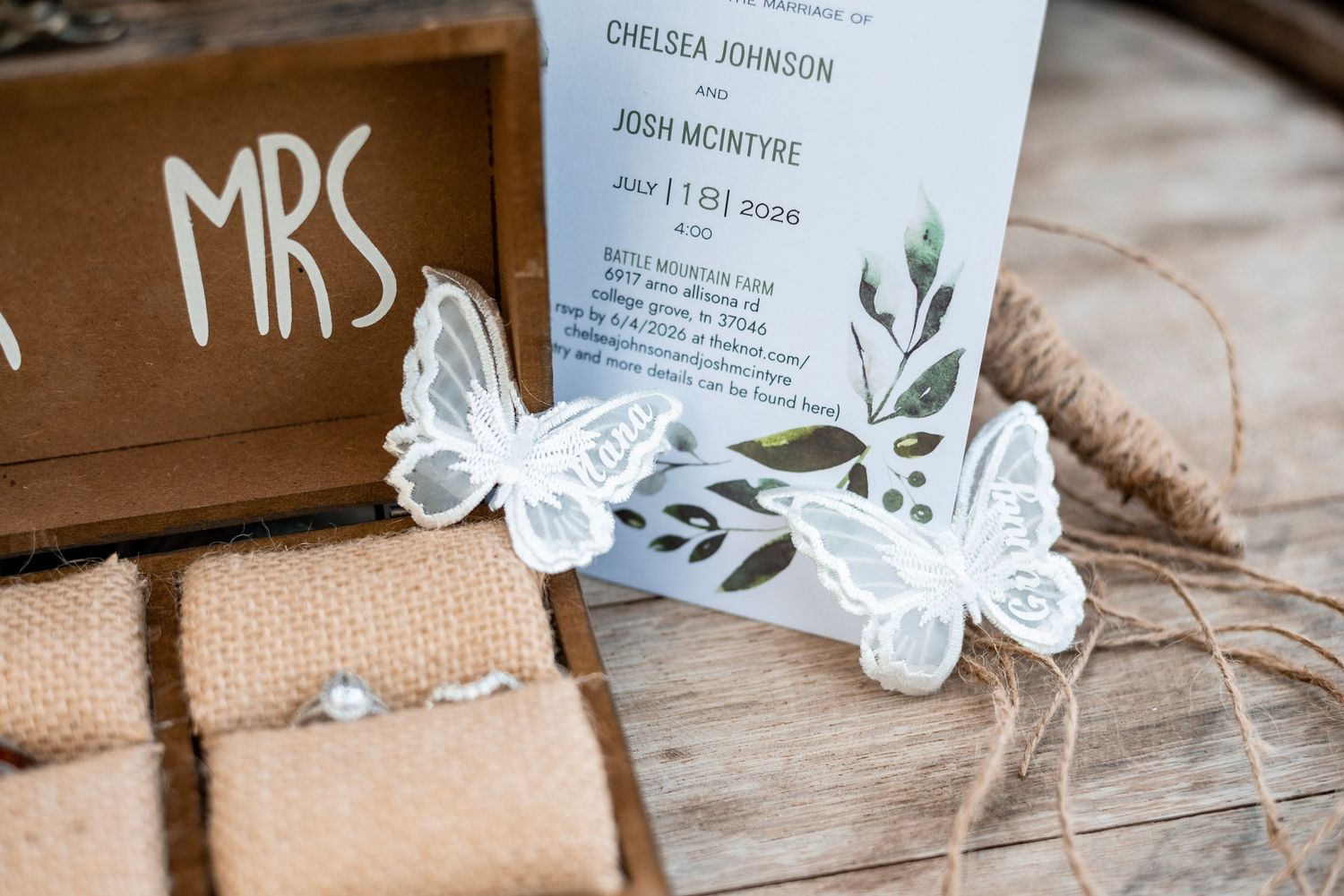 Elegant wedding invitation for Chelsea Johnson and Josh McIntyre with lace butterfly decorations and greenery on rustic wood.