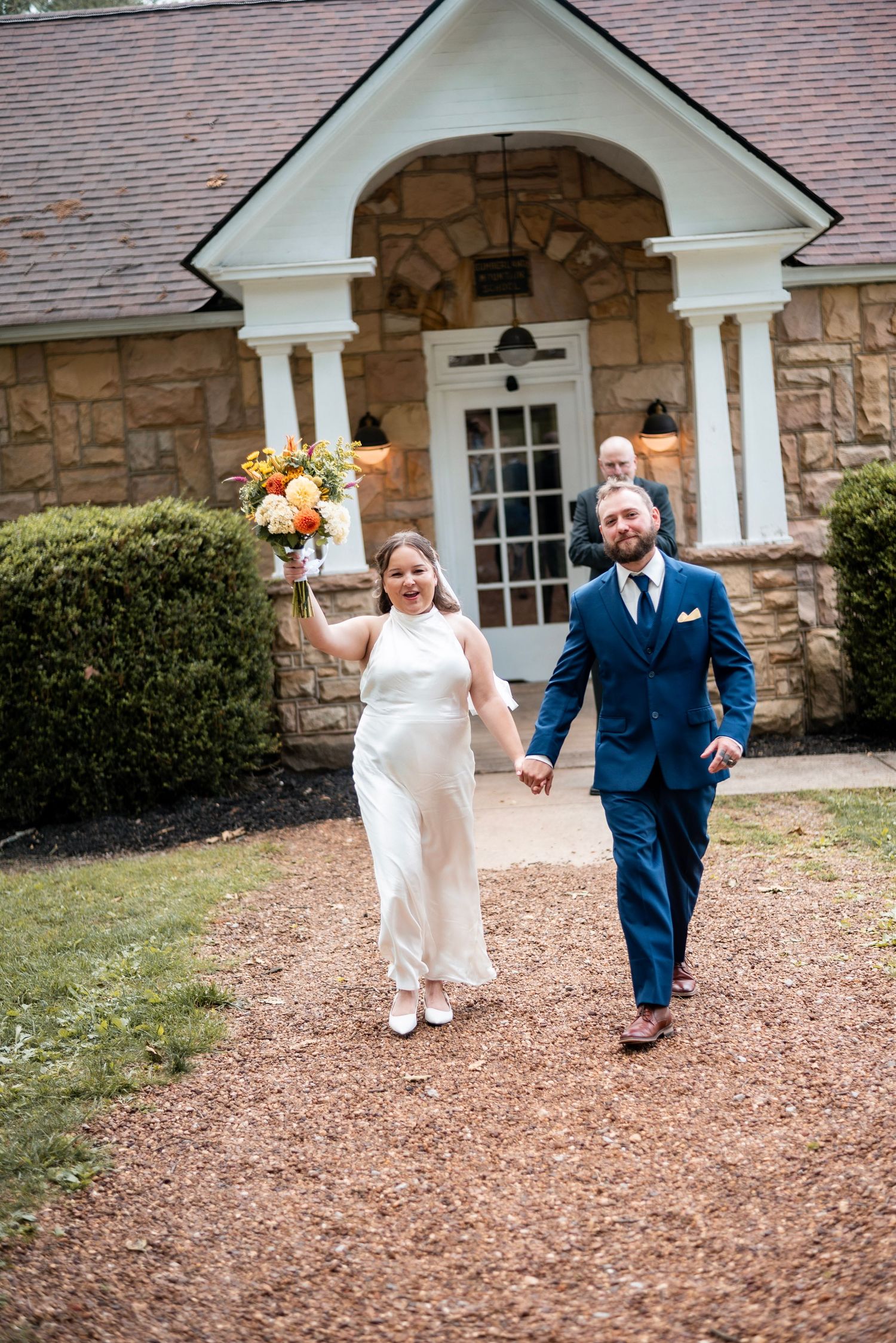 Bride and groom walk hand-in-hand from stone chapel, bride holding colorful bouquet, groom in navy suit.