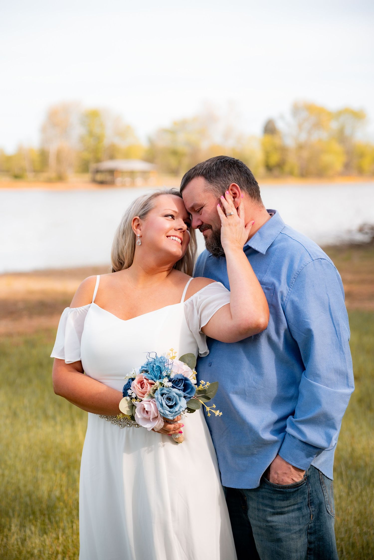 Couple sharing a romantic moment outdoors near a lake, bride in white dress holding blue and pink bouquet.