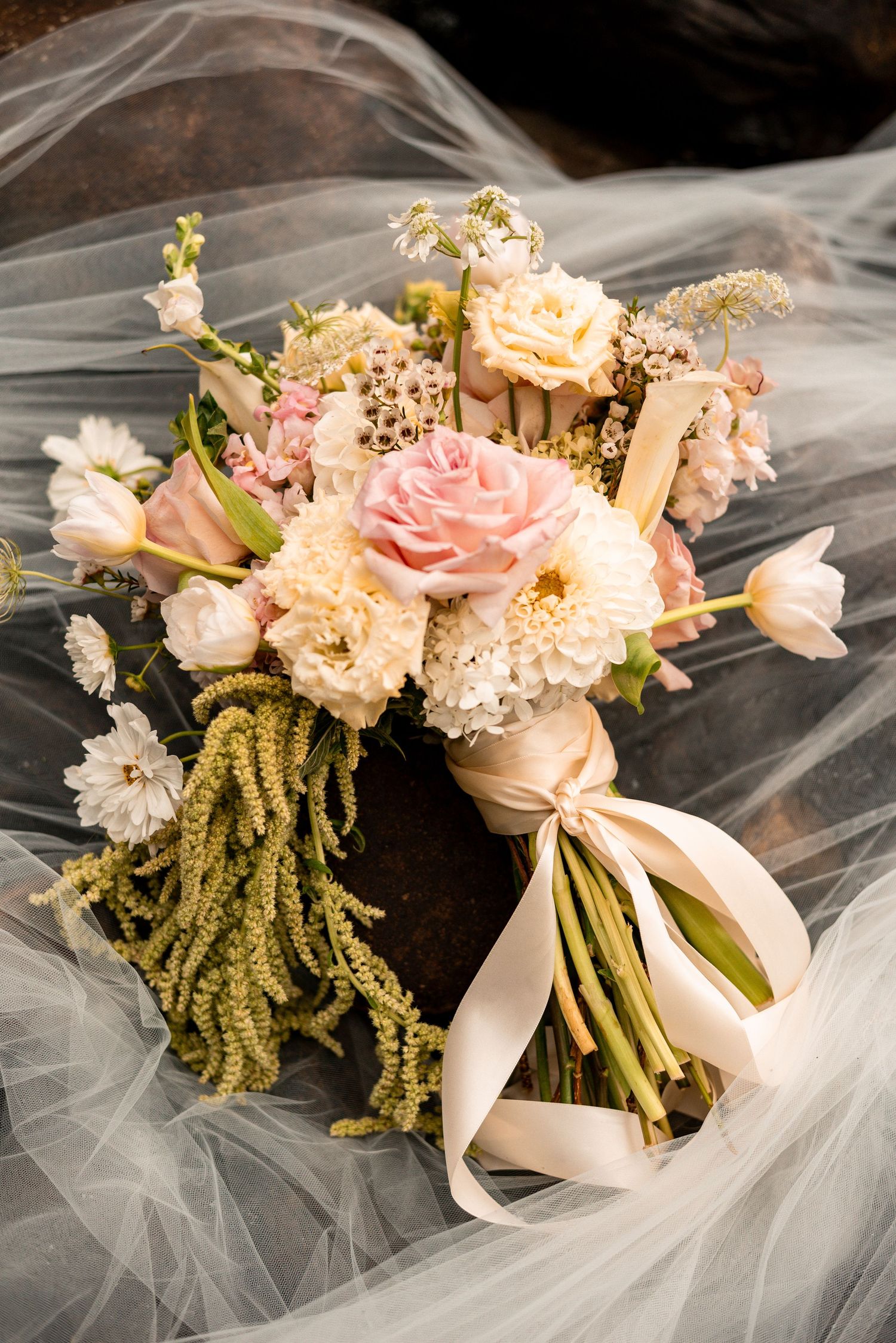 Elegant bridal bouquet with pink roses, cream dahlias, and trailing greenery tied with satin ribbon on sheer veil.