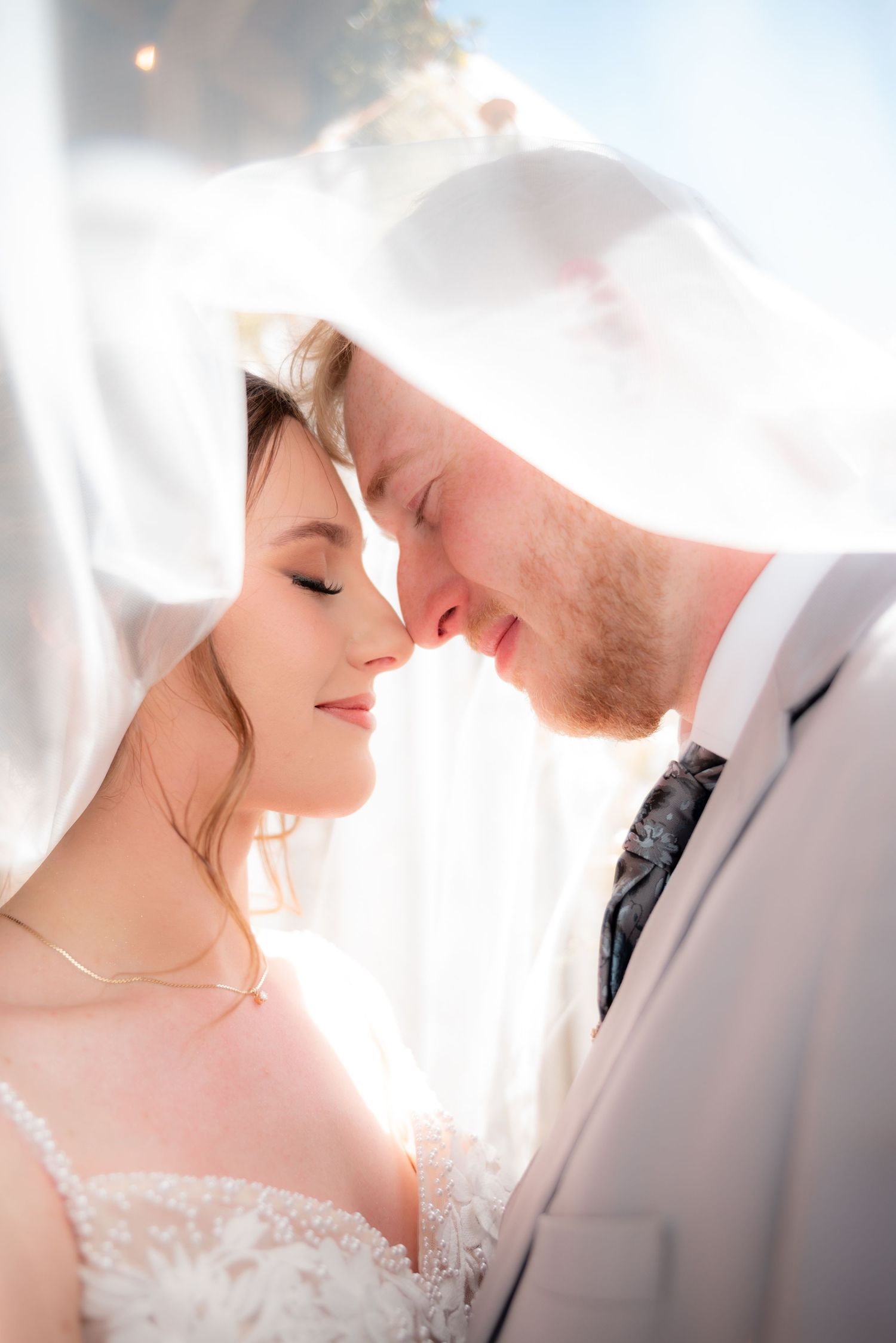 Bride and groom share an intimate forehead-to-forehead moment under a flowing white veil on their wedding day.