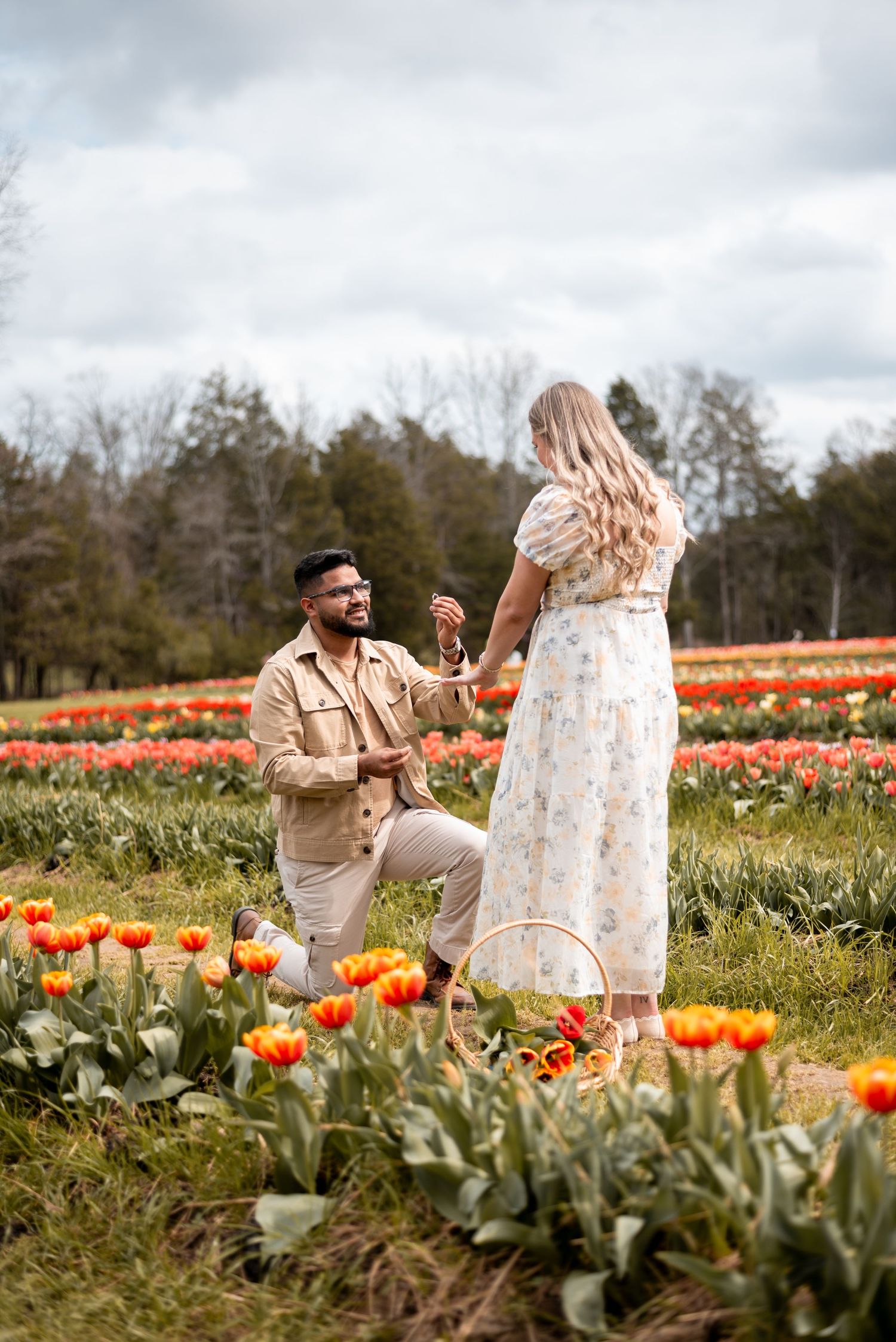 Man proposing to woman in a colorful tulip field, both smiling, with red and orange flowers surrounding them.