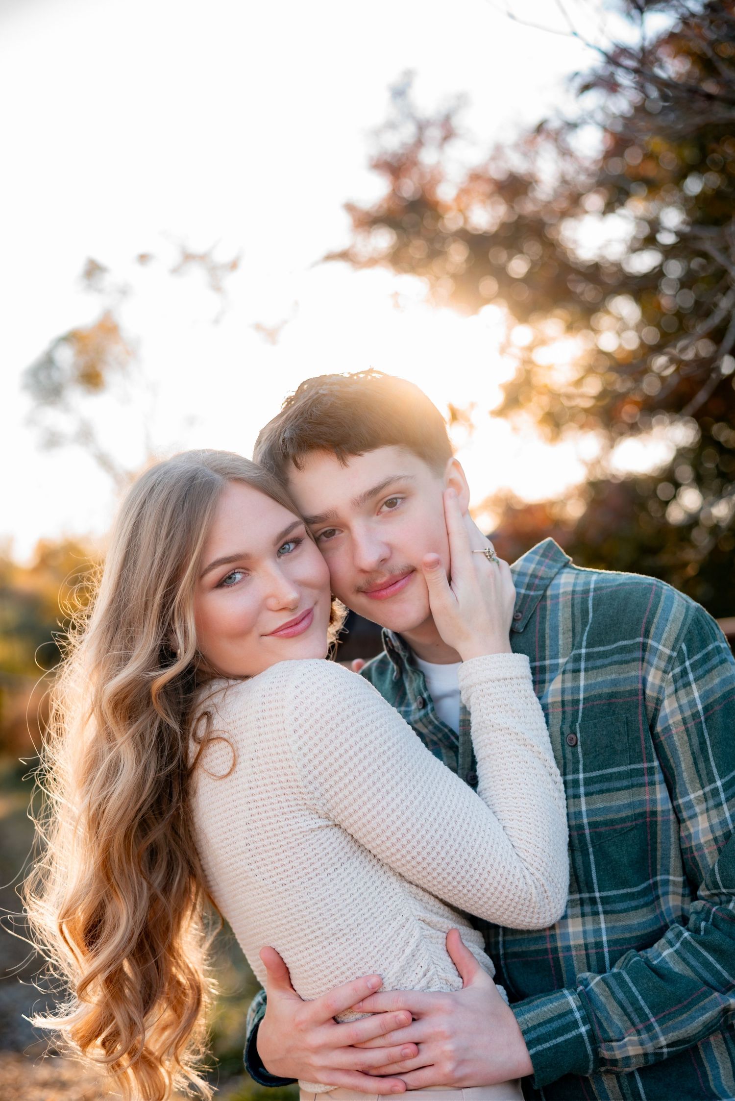 A couple in white sweater and plaid shirt embraces during a sunset photoshoot.