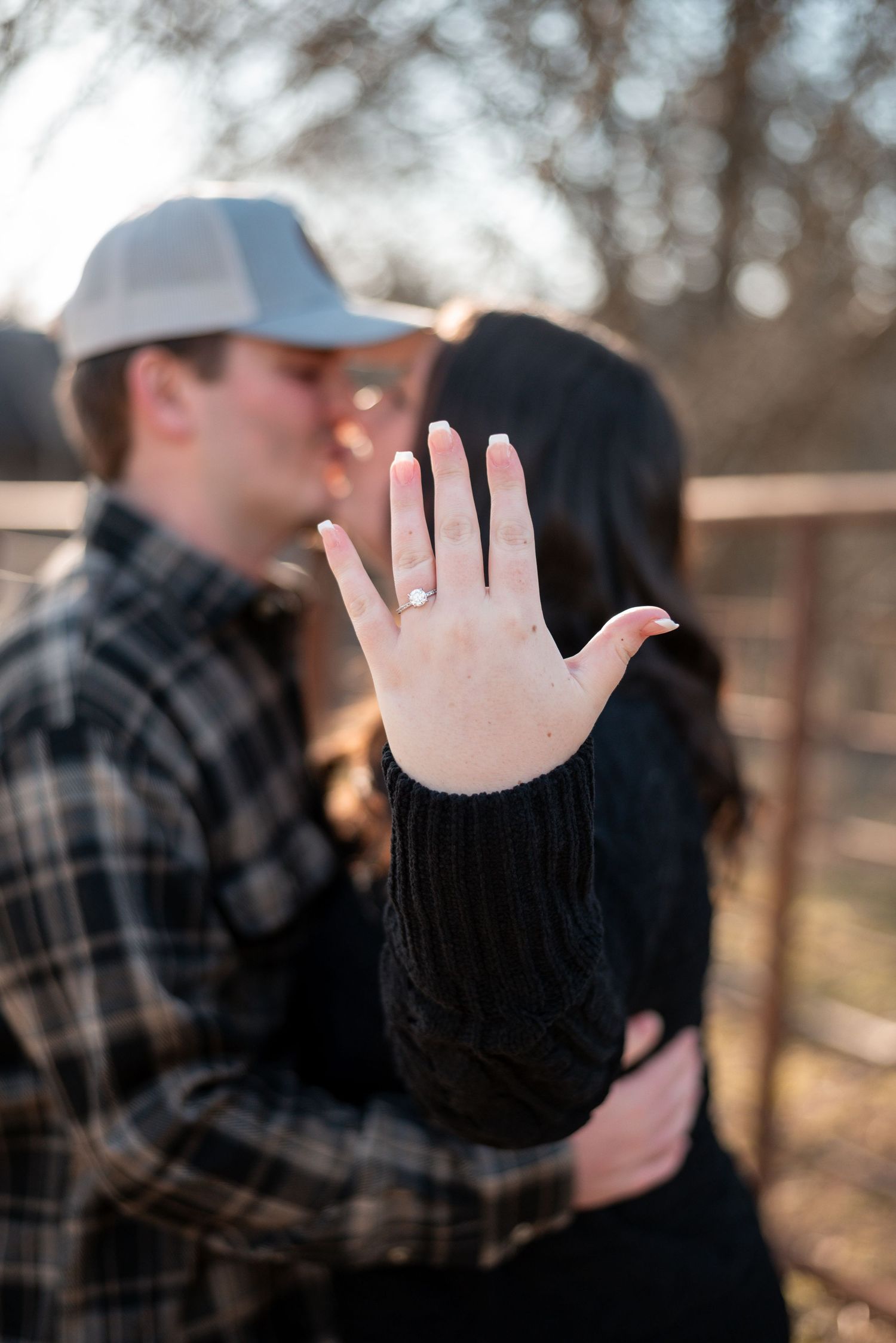 Hand showing off new engagement ring with couple embracing in soft focus background.