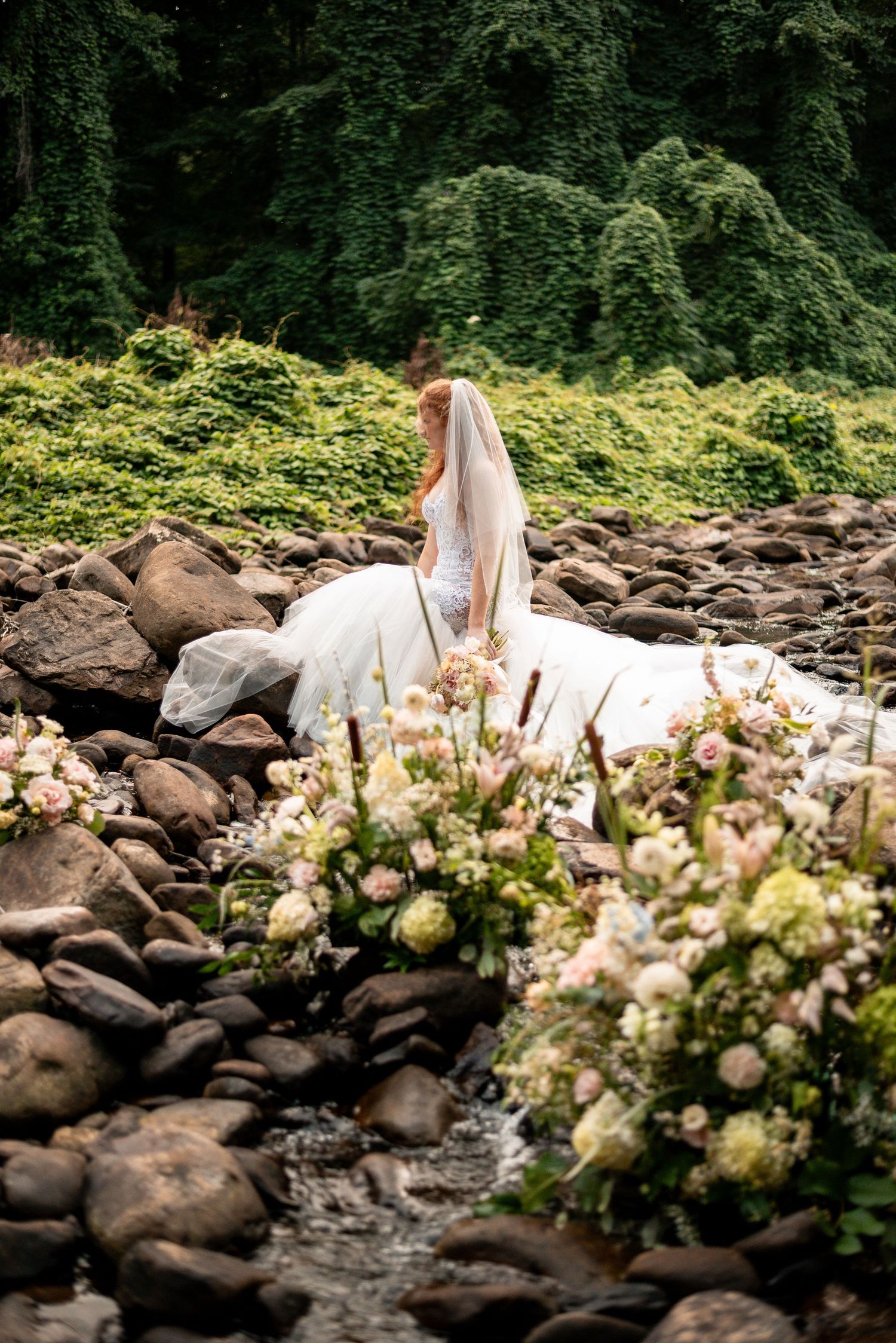 Bride in white gown and veil sits on rocky riverbed surrounded by lush greenery and elegant floral arrangements.