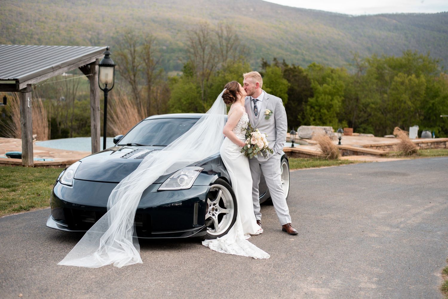 Bride and groom kissing beside a black sports car with mountain views at their outdoor wedding venue.