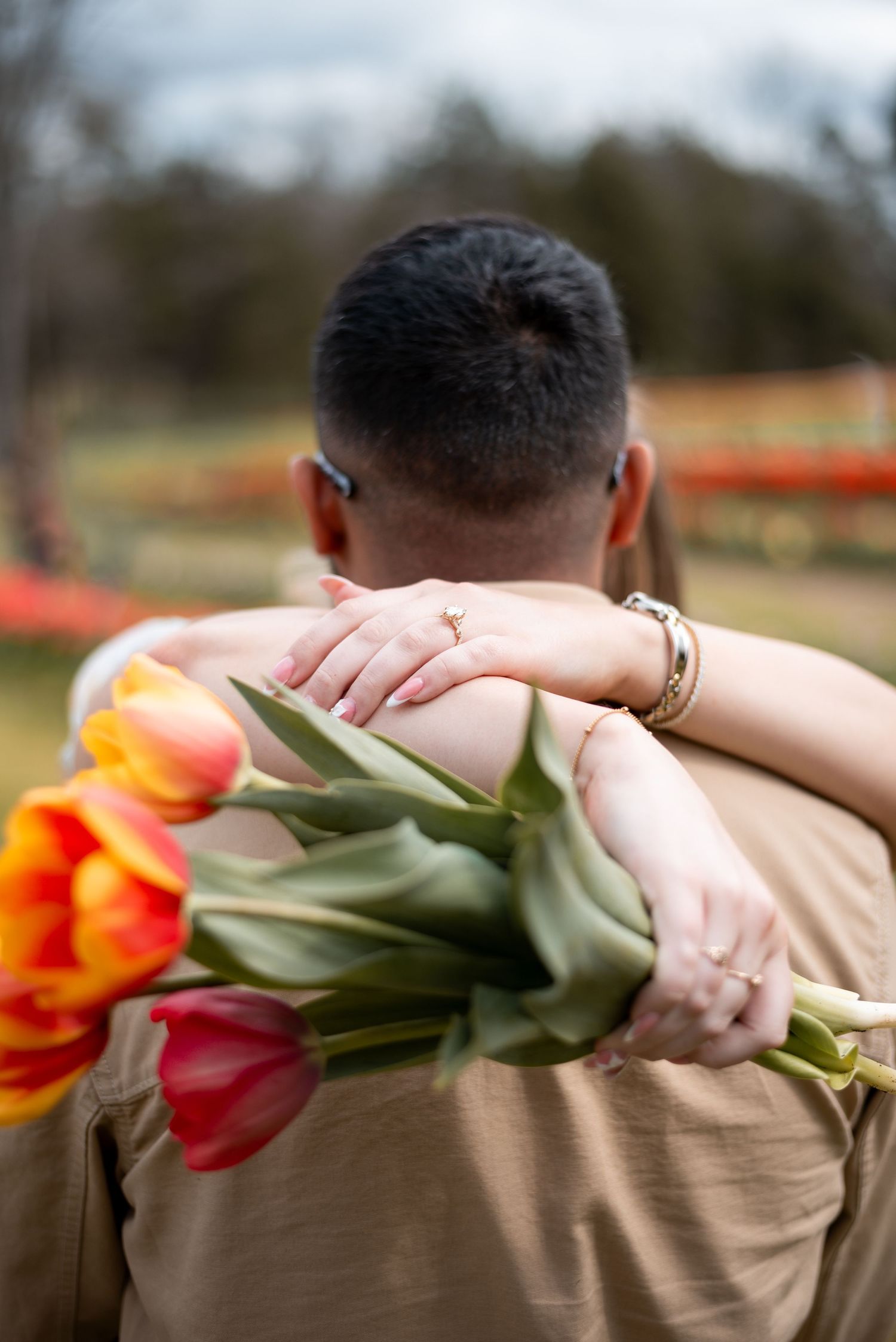 Couple embracing outdoors, woman holding vibrant red and orange tulip bouquet, viewed from behind in a romantic setting.