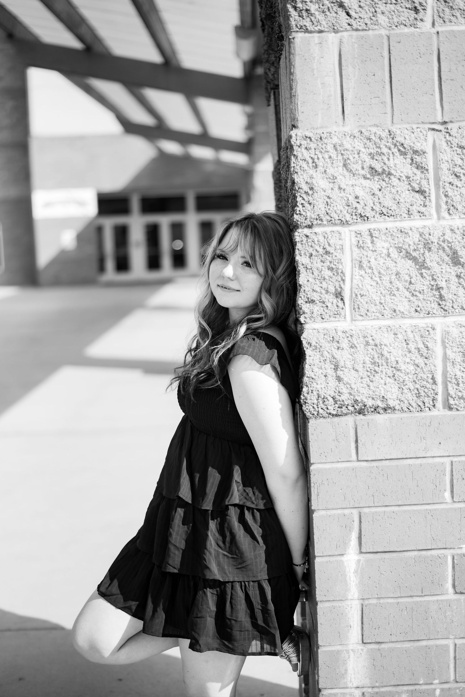 Black and white portrait of a young woman in a dress leaning against a brick wall outdoors.