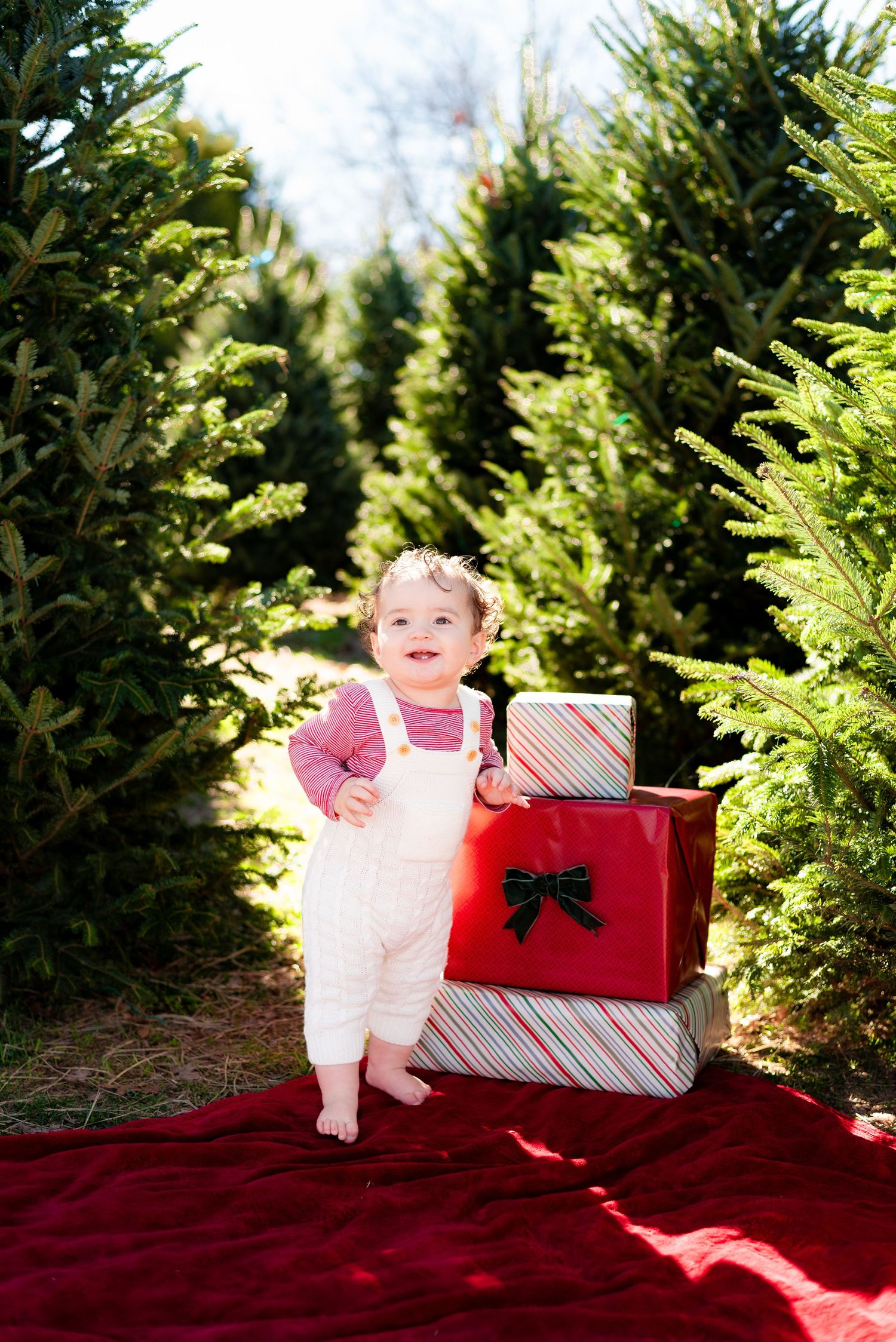 Baby in white outfit sitting among Christmas presents on red blanket at tree farm.