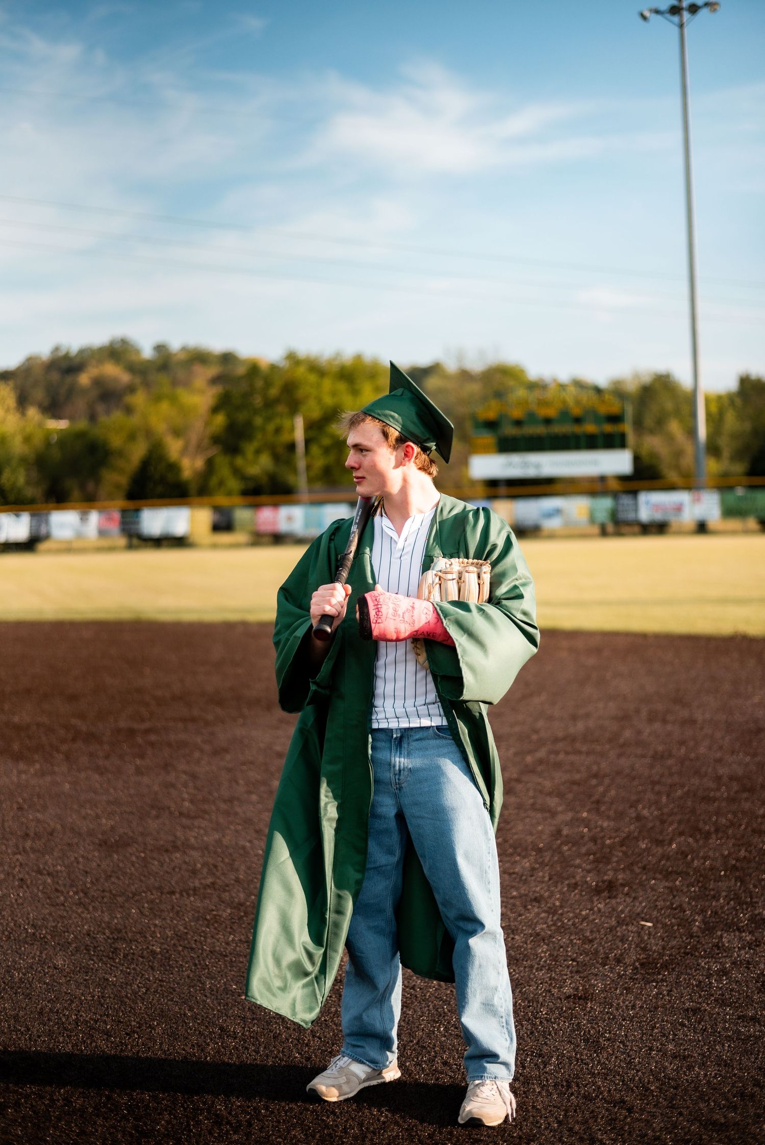 Young man in green graduation cap and gown standing on a sports field holding diploma on a sunny day.