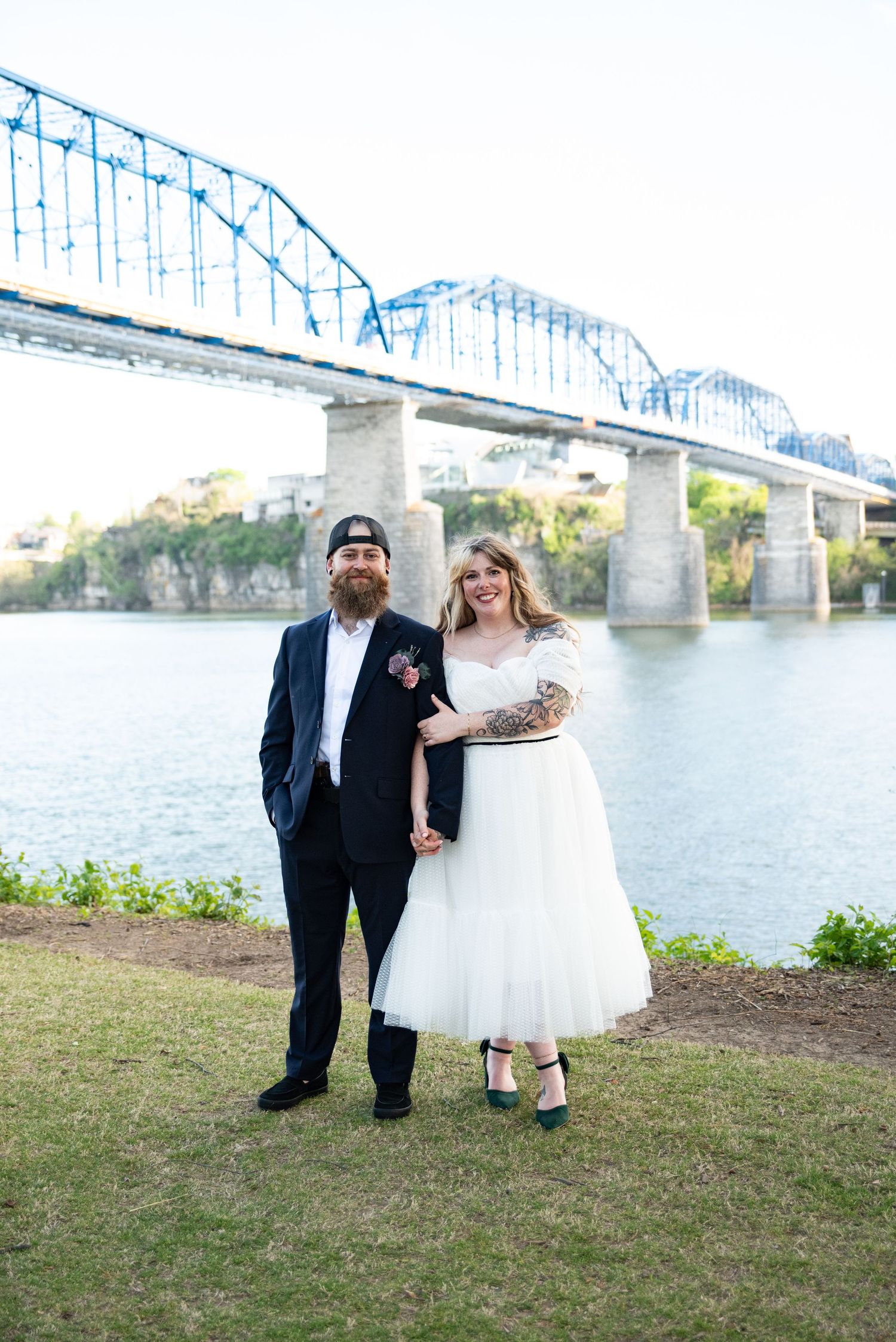 Bride and groom pose by a river with a blue steel bridge backdrop on their wedding day.