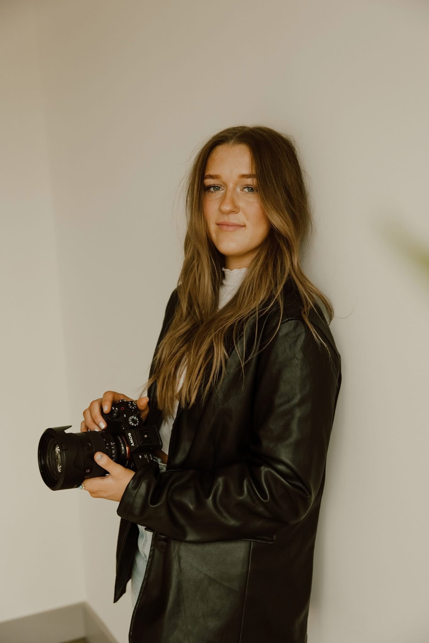 Young woman in black leather jacket holding a DSLR camera, posing against a white wall with long brown hair.