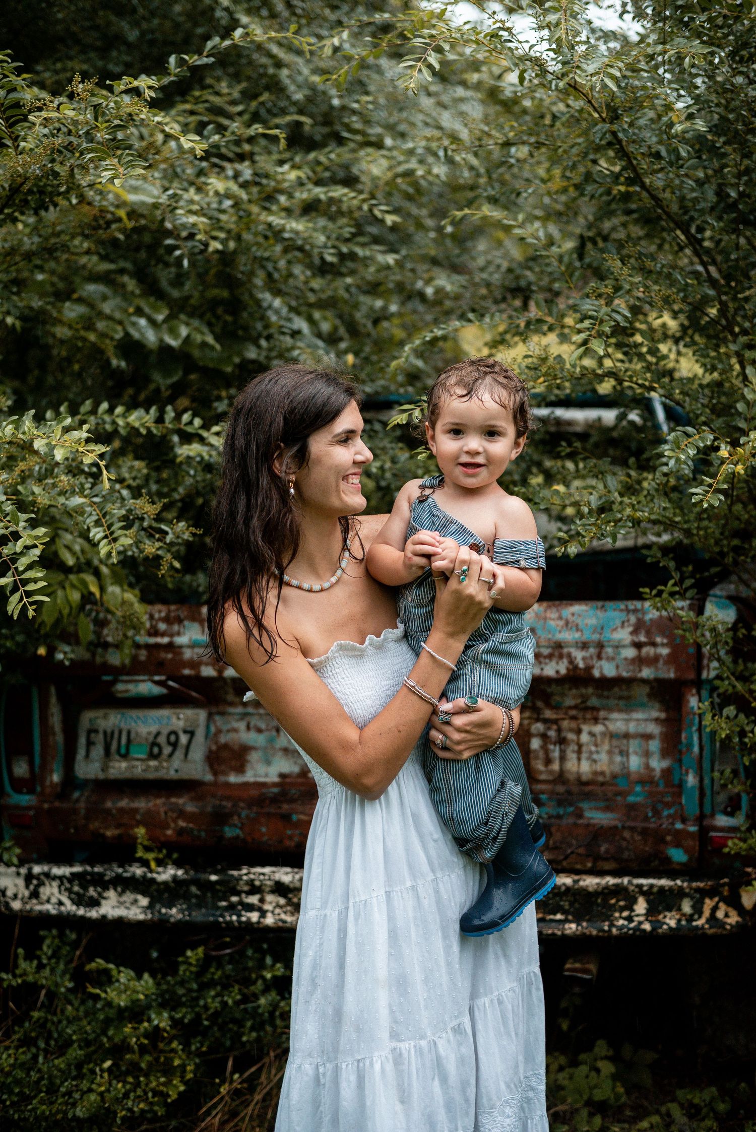 A smiling woman in a white dress holds a happy toddler in denim overalls outdoors near rustic greenery.