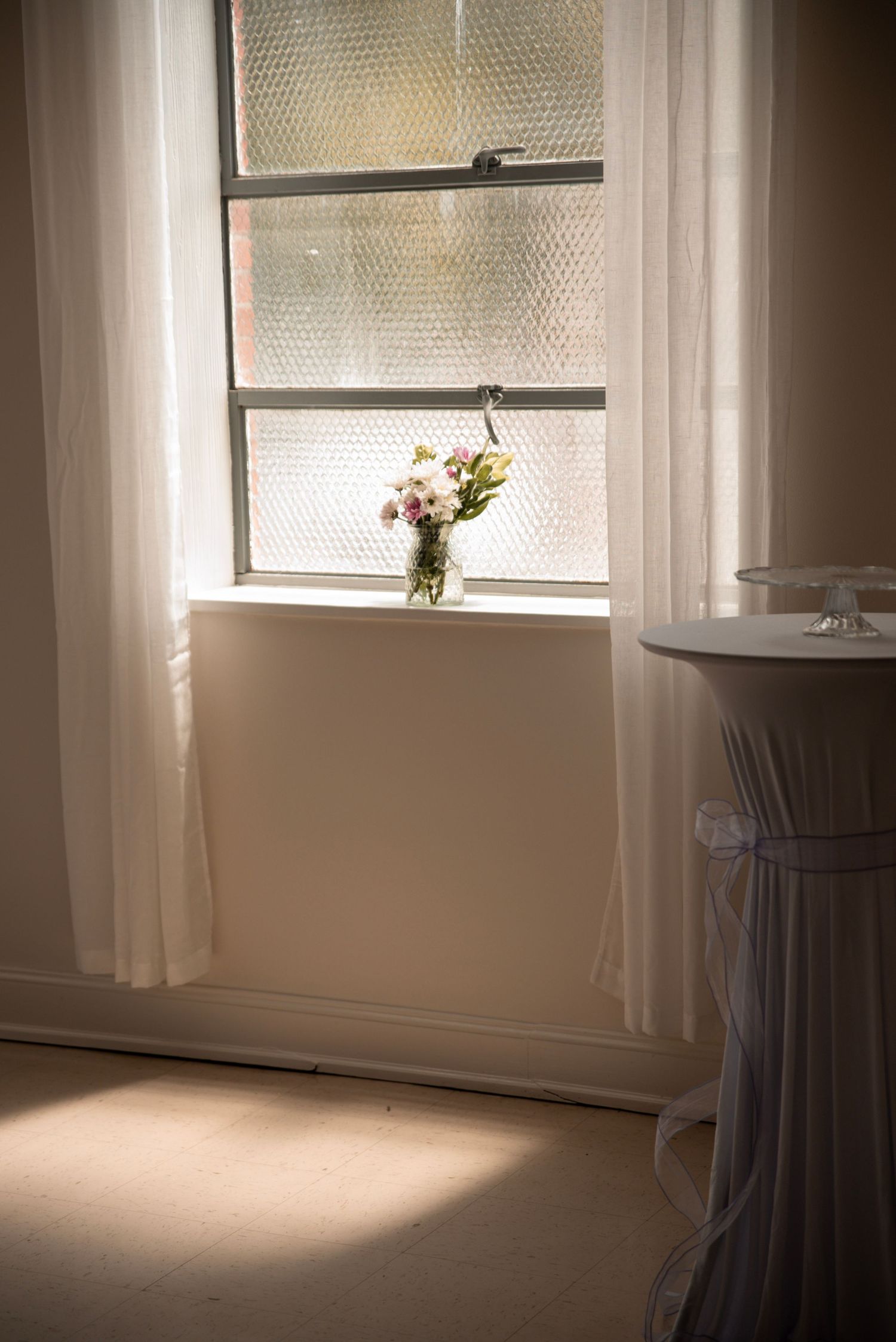 Sunlit window with sheer white curtains and a small floral arrangement in a glass vase on the sill.