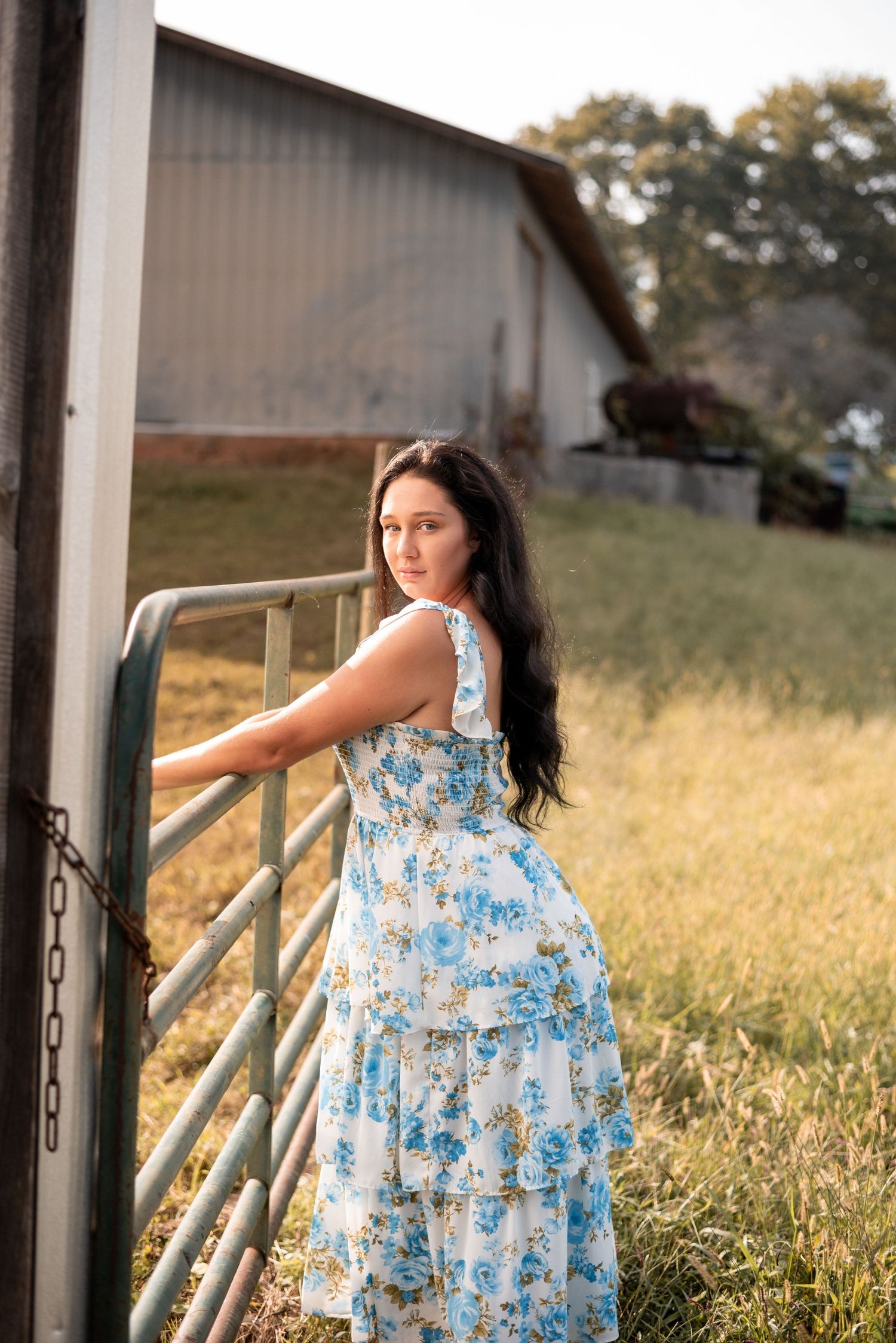 A person in a flowing blue dress poses by a metal gate near a rural barn at sunset.