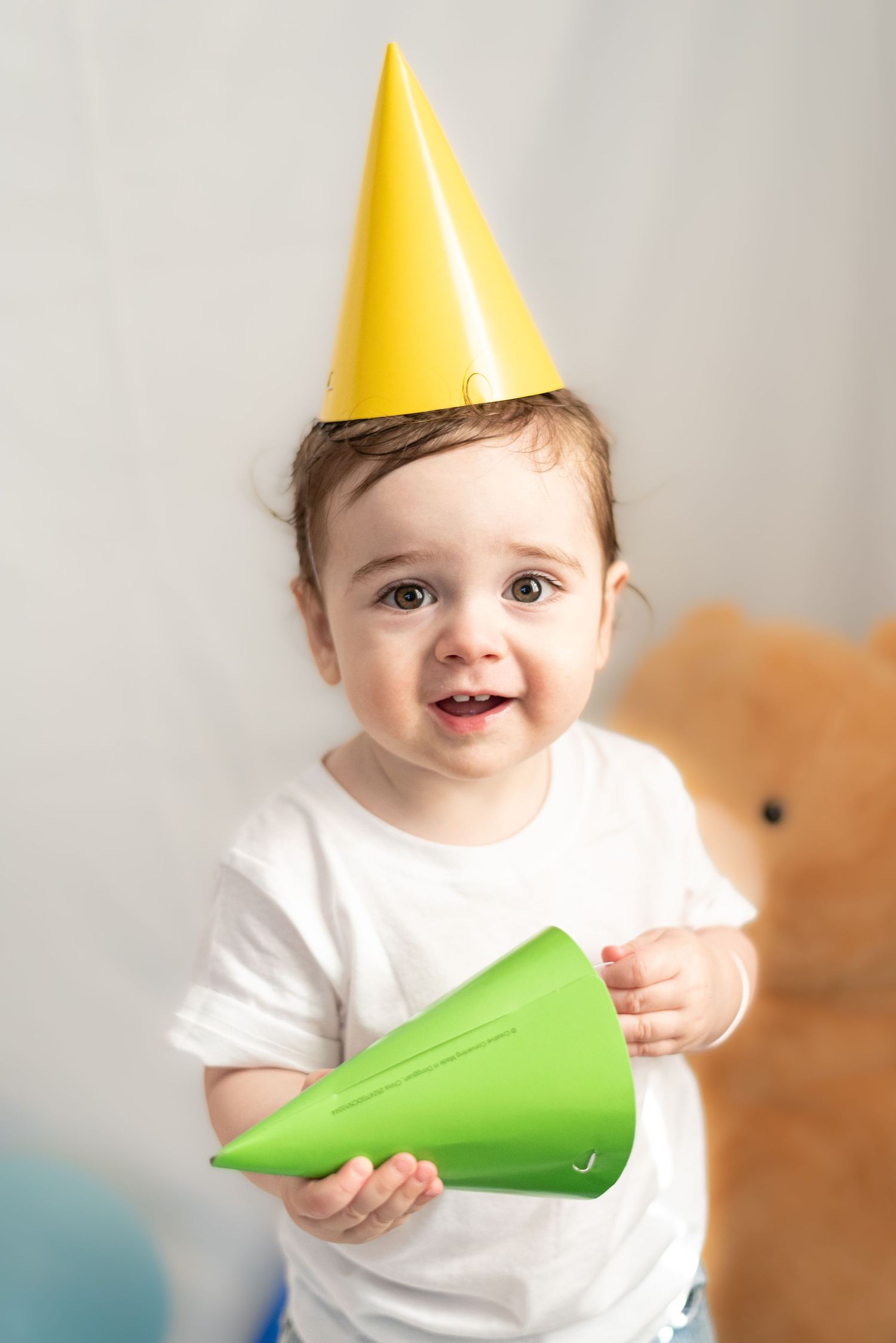 A toddler wearing a yellow party hat and white shirt holds a green party horn.