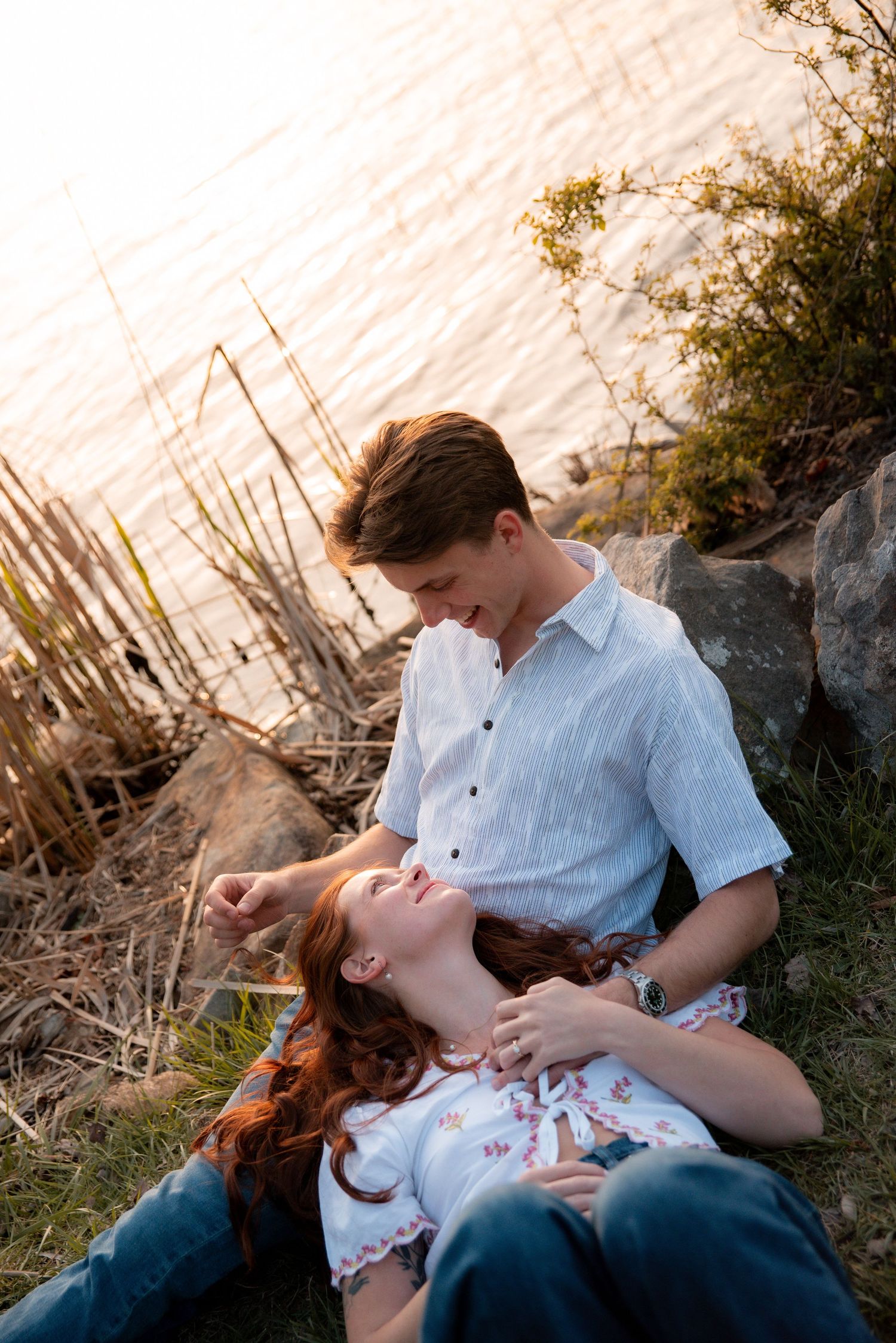 Couple relaxing by a sunlit lake shore, woman lying in mans lap smiling, golden hour romantic outdoor portrait.