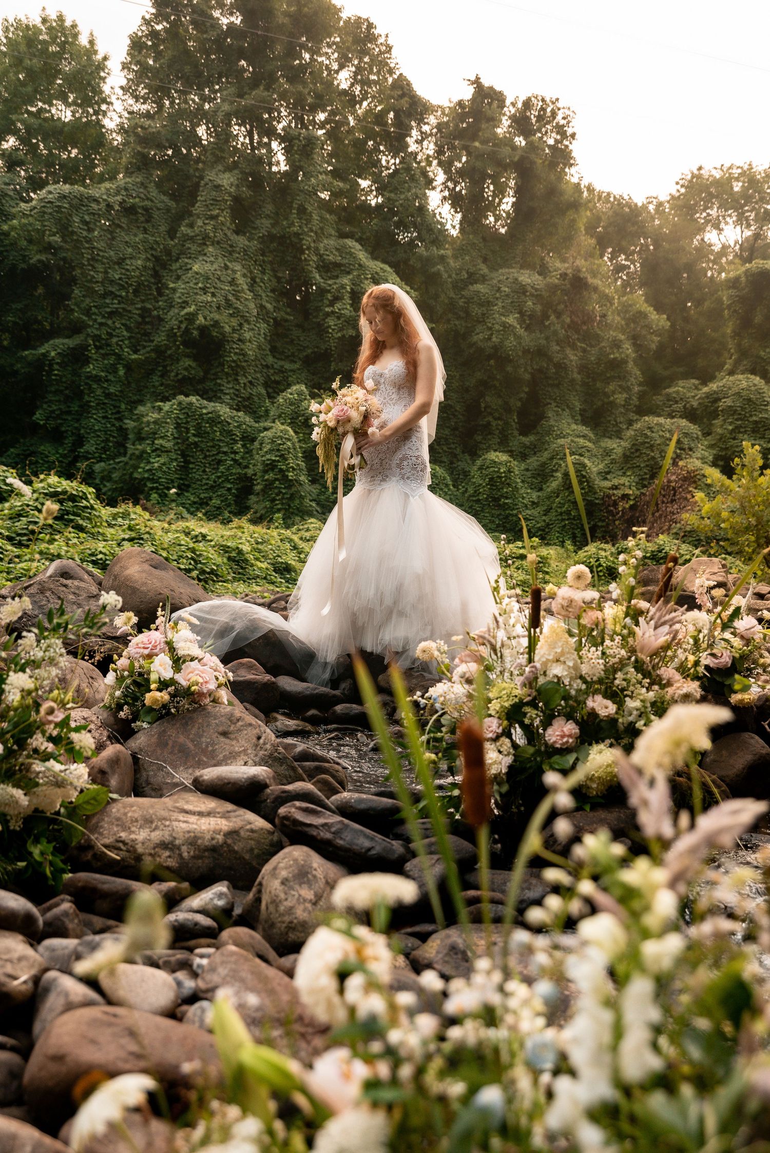 Bride in white mermaid gown holding bouquet stands among rocks and wildflowers in a lush green garden setting.