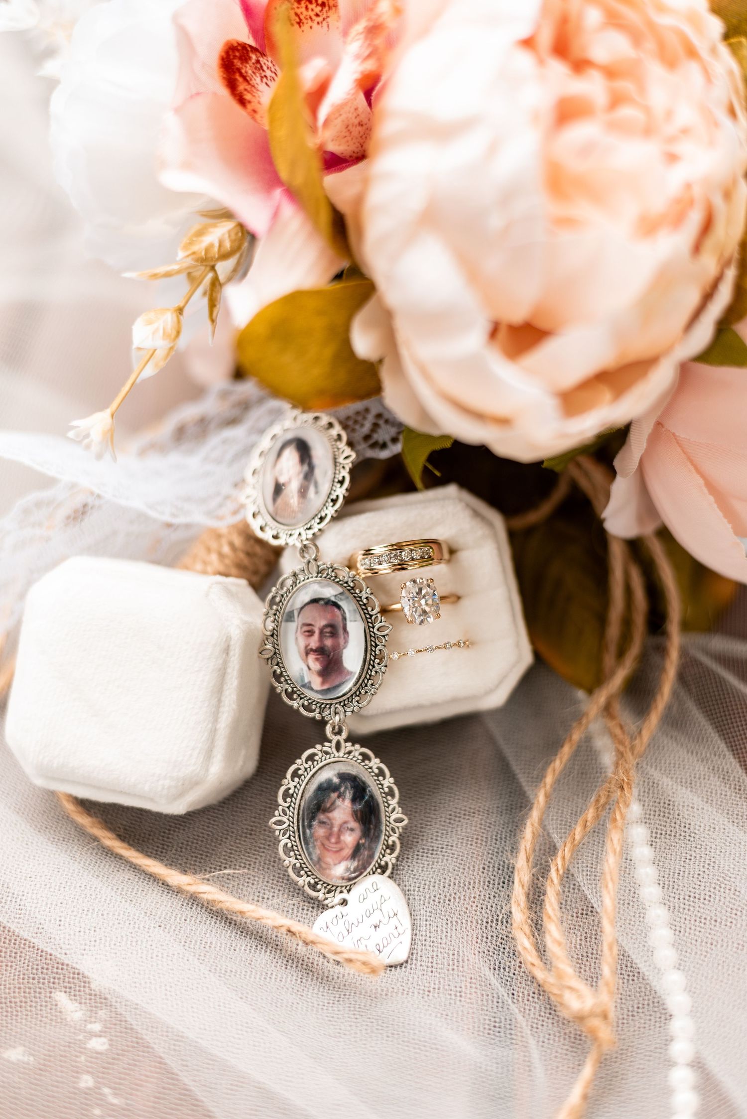 Wedding bouquet with memorial photo charms, diamond engagement ring in velvet box, and soft pink roses on tulle fabric.