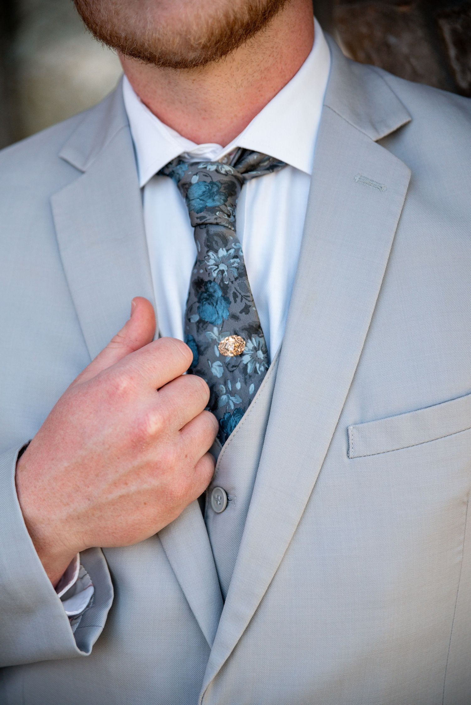 Man in light gray suit adjusting a blue floral tie with a decorative tie pin.