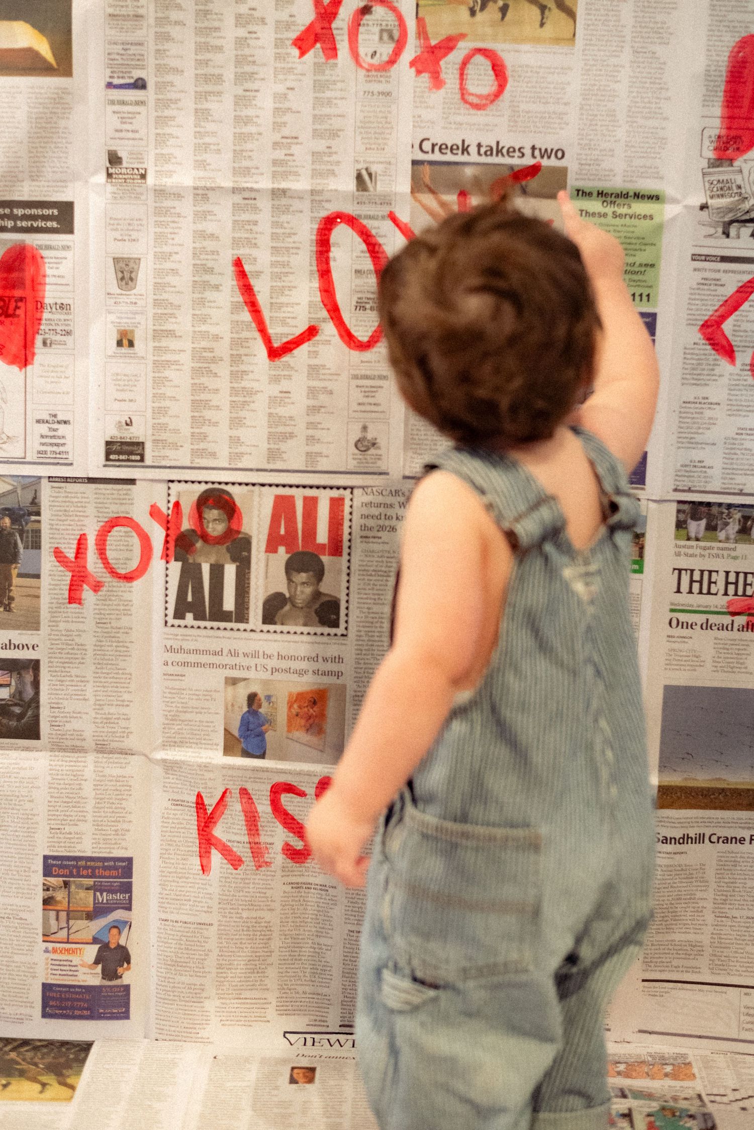 A toddler in overalls reaches up to newspaper-covered walls with red writing.
