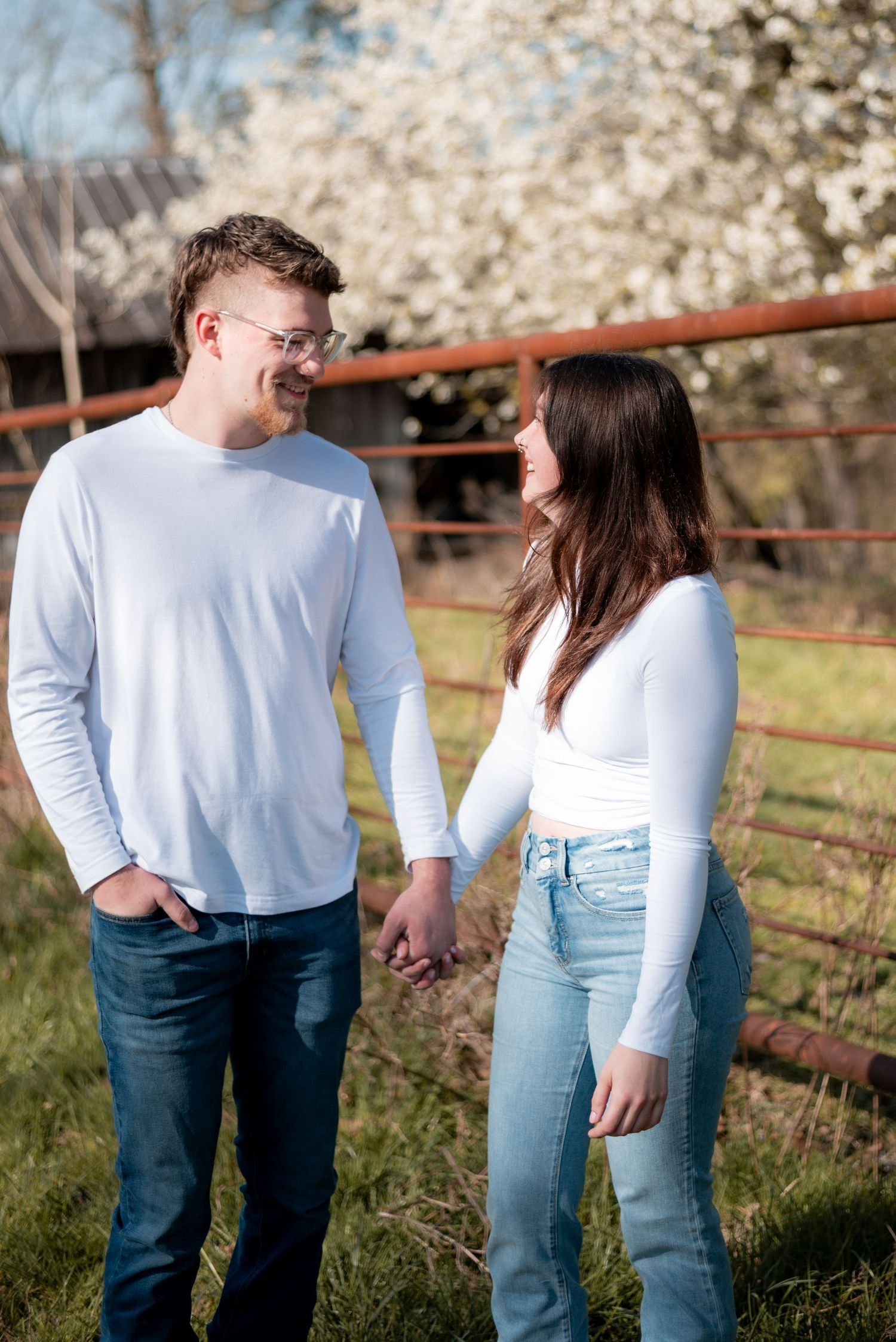 A smiling couple in matching white tops and jeans hold hands outdoors near a rustic fence with blooming spring trees.