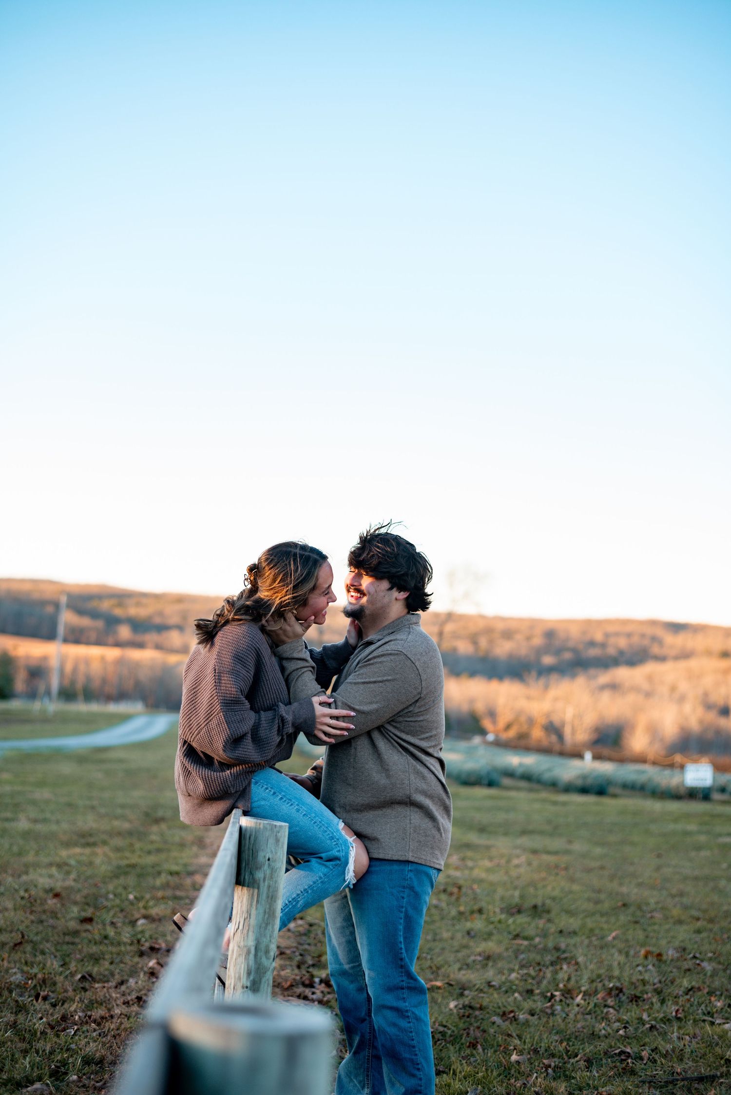 Couple sharing a tender moment while leaning against a wooden fence in a rural field.