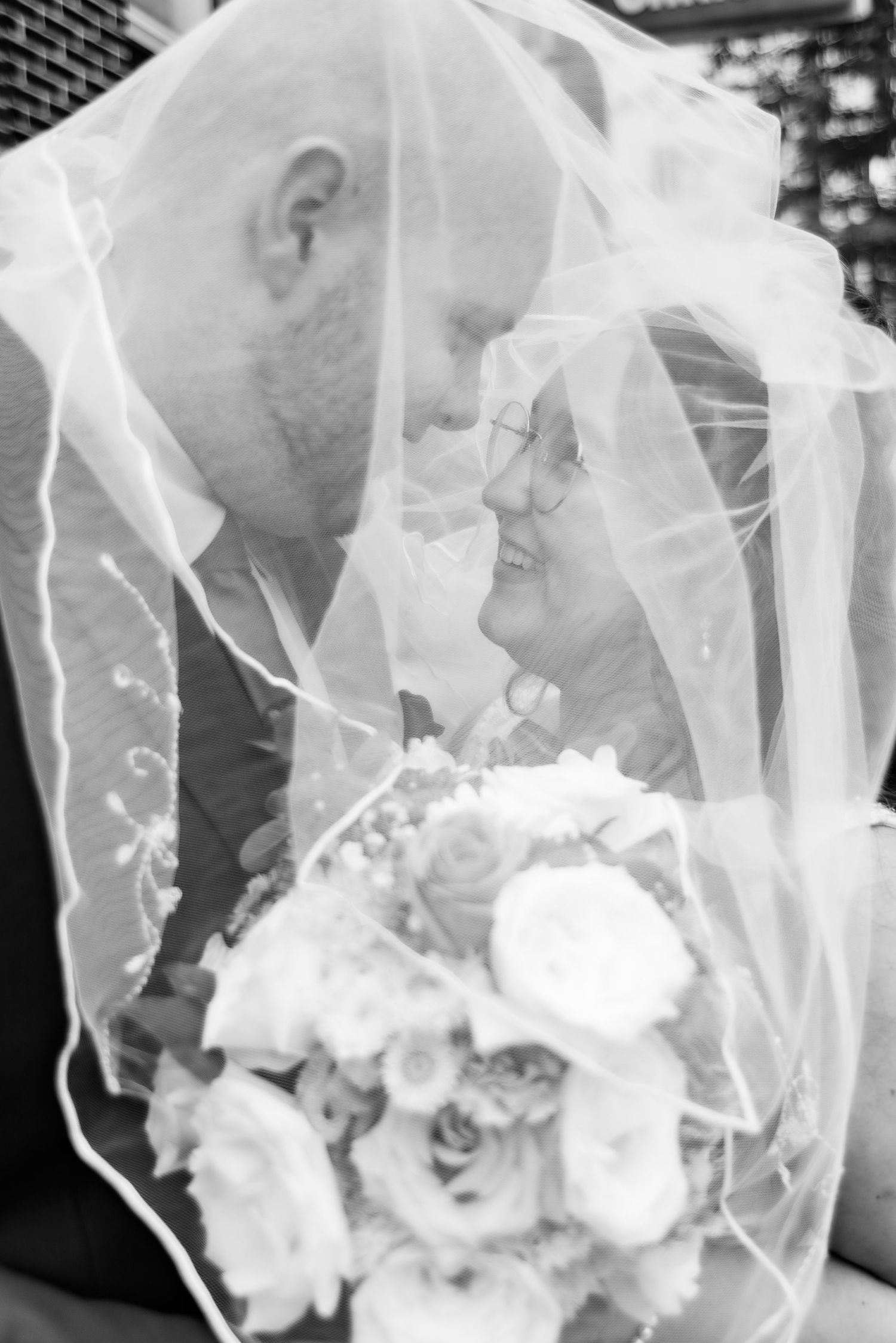 Black and white wedding photo of bride and groom sharing an intimate moment under a veil, bride holding a bouquet.