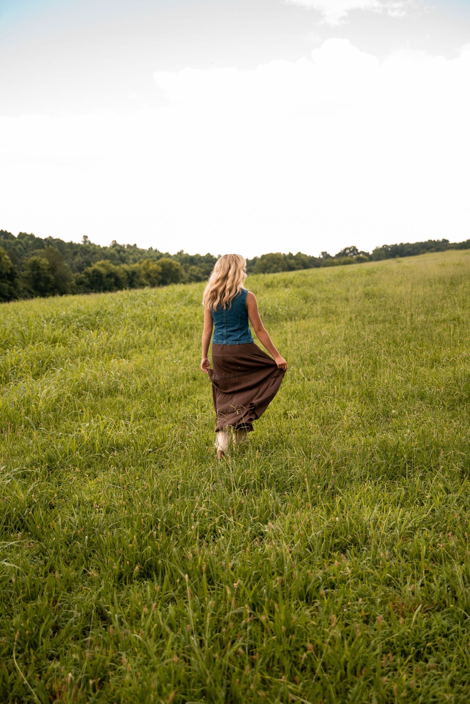 A blonde woman in a teal top and brown maxi skirt walks barefoot through a lush green meadow.