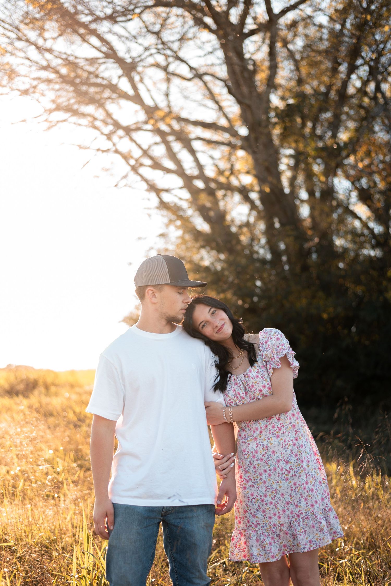 A couple walks together in a sunlit field during golden hour with trees in the background.